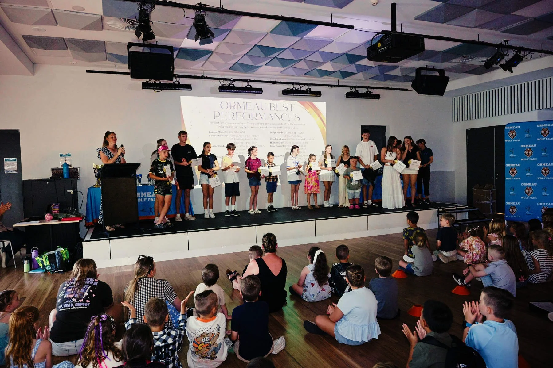 Children and teenagers standing on stage during an award ceremony, some holding certificates, with an audience of kids sitting on the floor watching. A woman in yellow and a woman in white are among the presenters. A large screen behind displays text