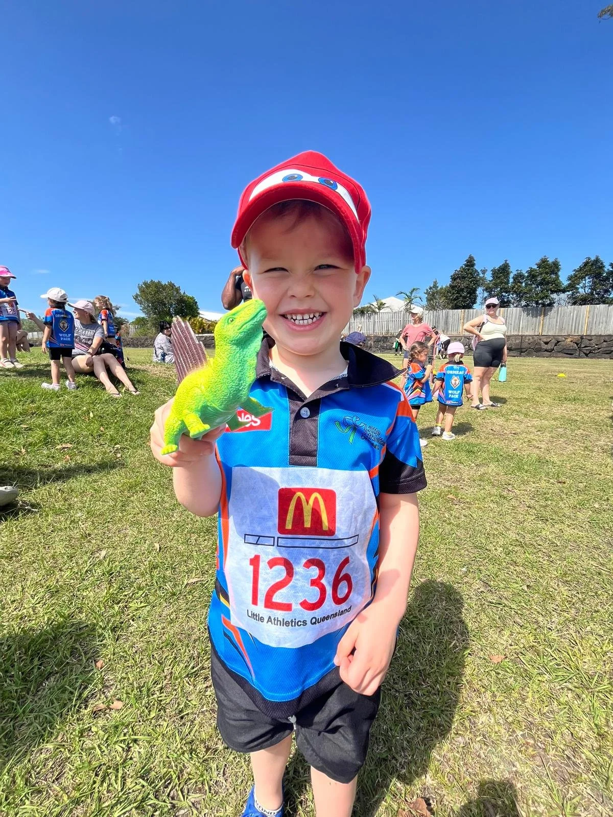 A young boy wearing a red hat and a blue sports jersey with a McDonald's logo and the number 1236, holding a green toy lizard, smiling outdoors on a sunny day with other children and adults in the background.