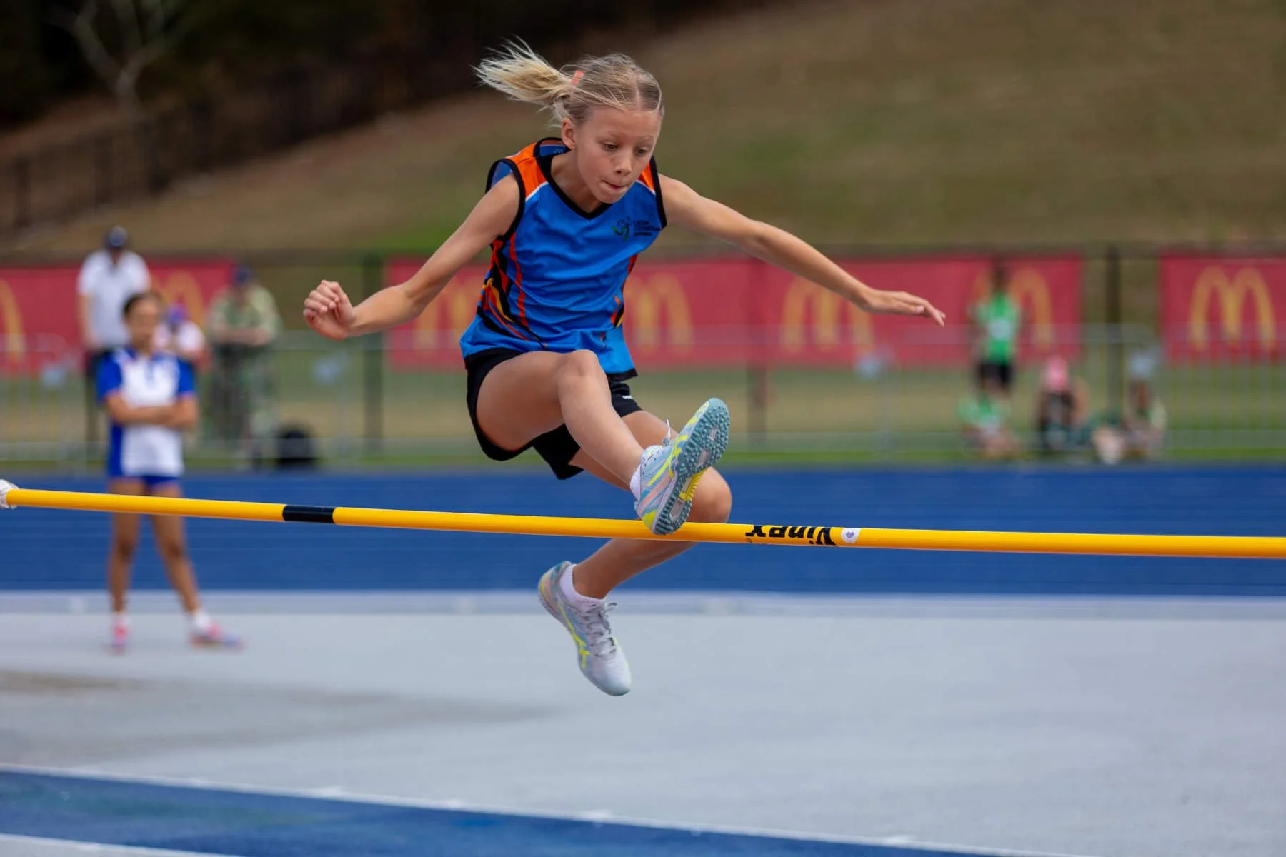 A young girl in athletic attire clearing a yellow hurdle on a track field with a blue running track and blurred spectators in the background.