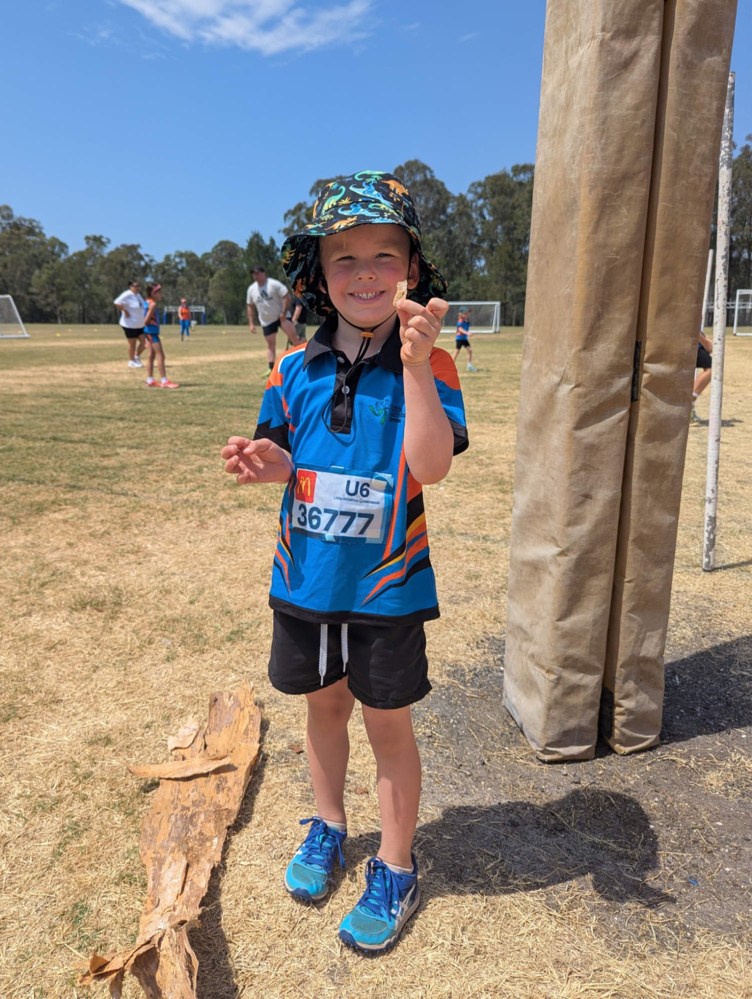 Young boy in athletic gear standing outdoors at a sports field, smiling and holding a shell, with a door frame to the right and other people in the background.