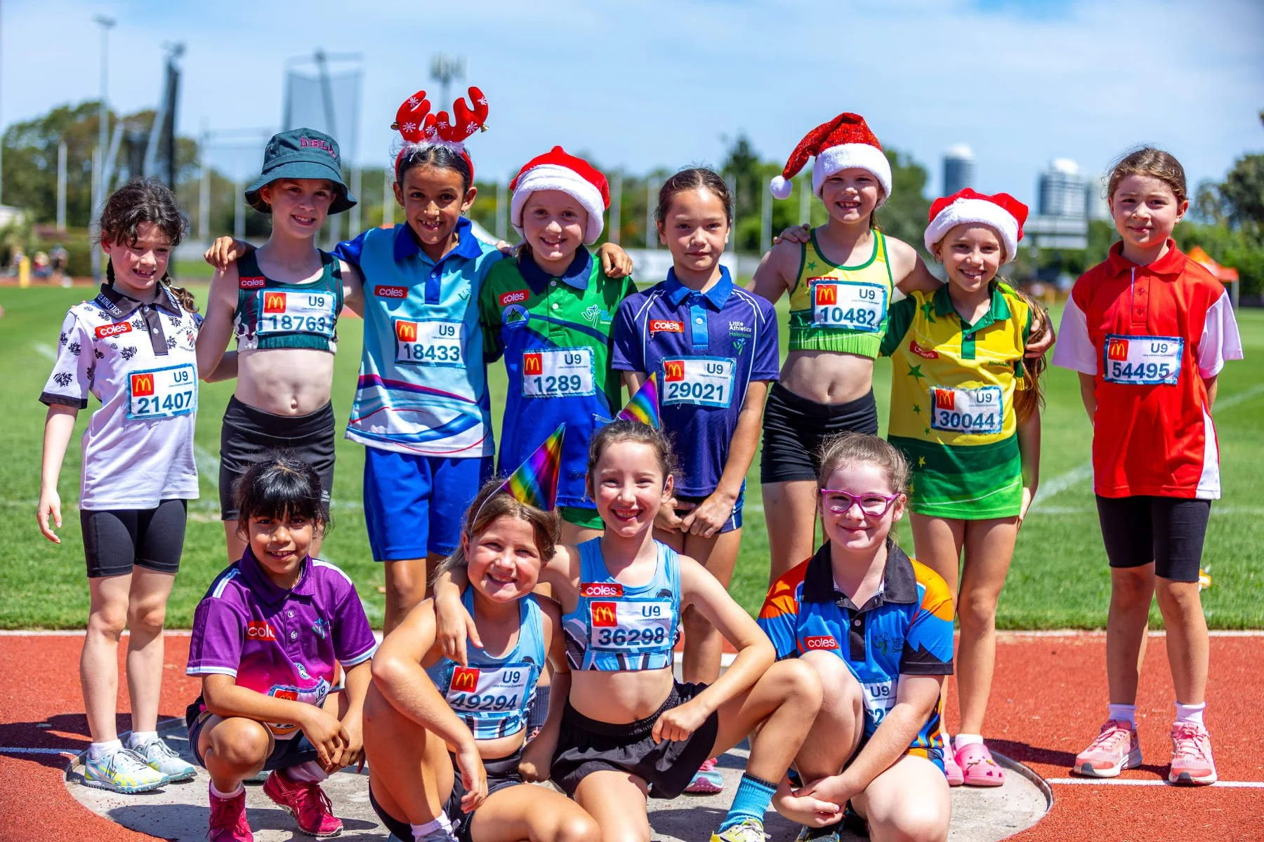 A group of young children in athletic clothing and Santa hats posing for a photo on a track field during a festive outdoor event.