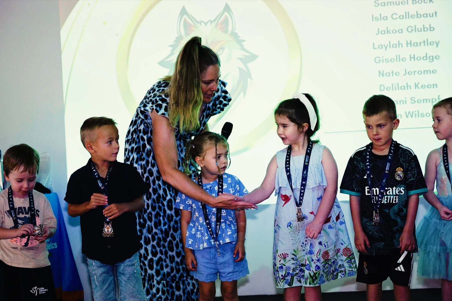 A group of children on stage receiving awards from a woman in a blue animal print dress, with a screen displaying a list of names in the background.