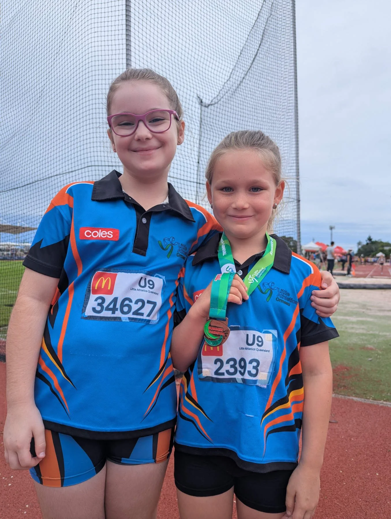 Two young girls in athletic uniforms standing on a sports field, one holding medals and smiling.
