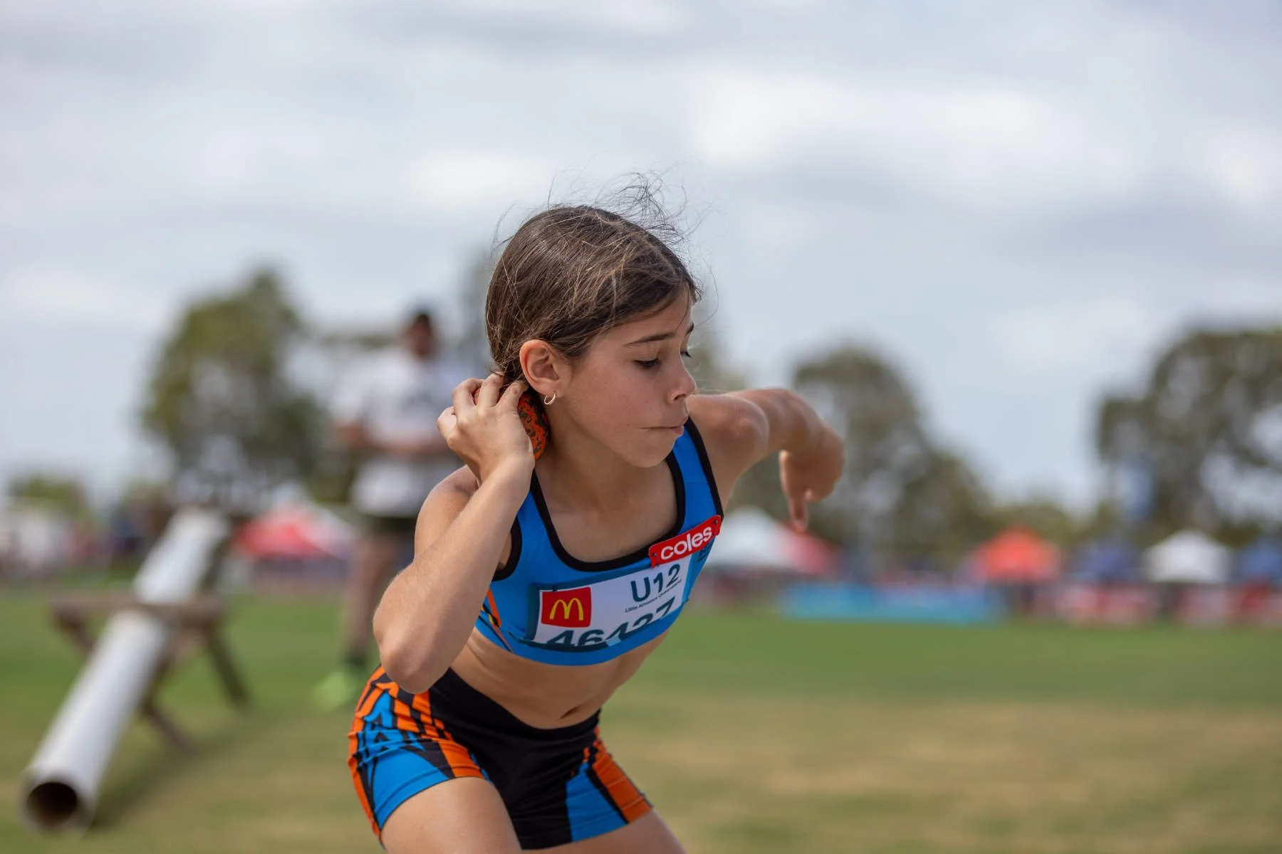 Young female athlete in blue and orange running uniform, leaning forward and preparing to race, outdoors on a cloudy day.