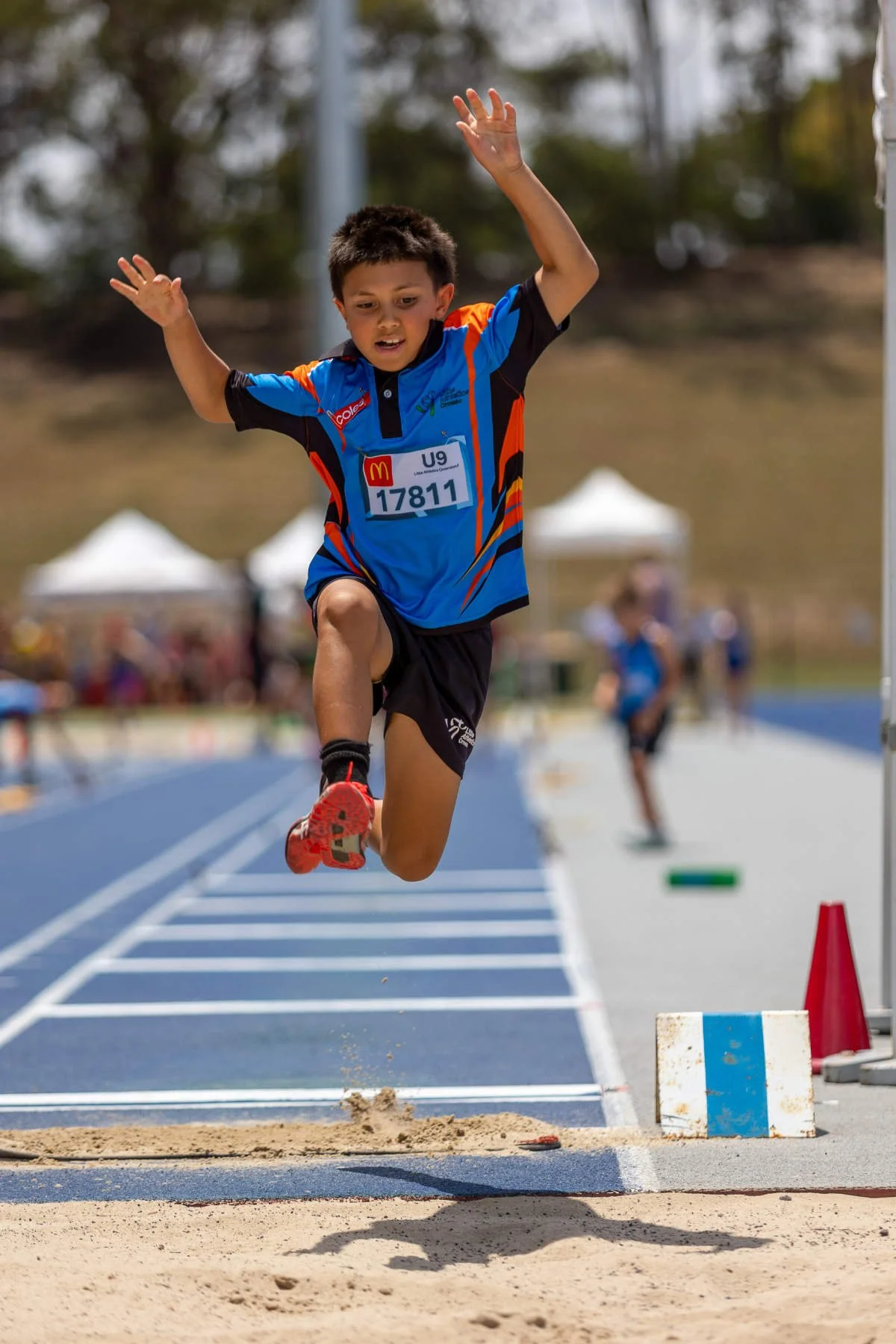 A young boy wearing a blue, orange, and black sports uniform and red running shoes is captured mid-air as he jumps into a sandpit during a track and field long jump event. He has a focused expression, and a race bib with the number 17811 is pinned to his shirt. The background shows a blue running track, other children, and tents in an outdoor athletic field.