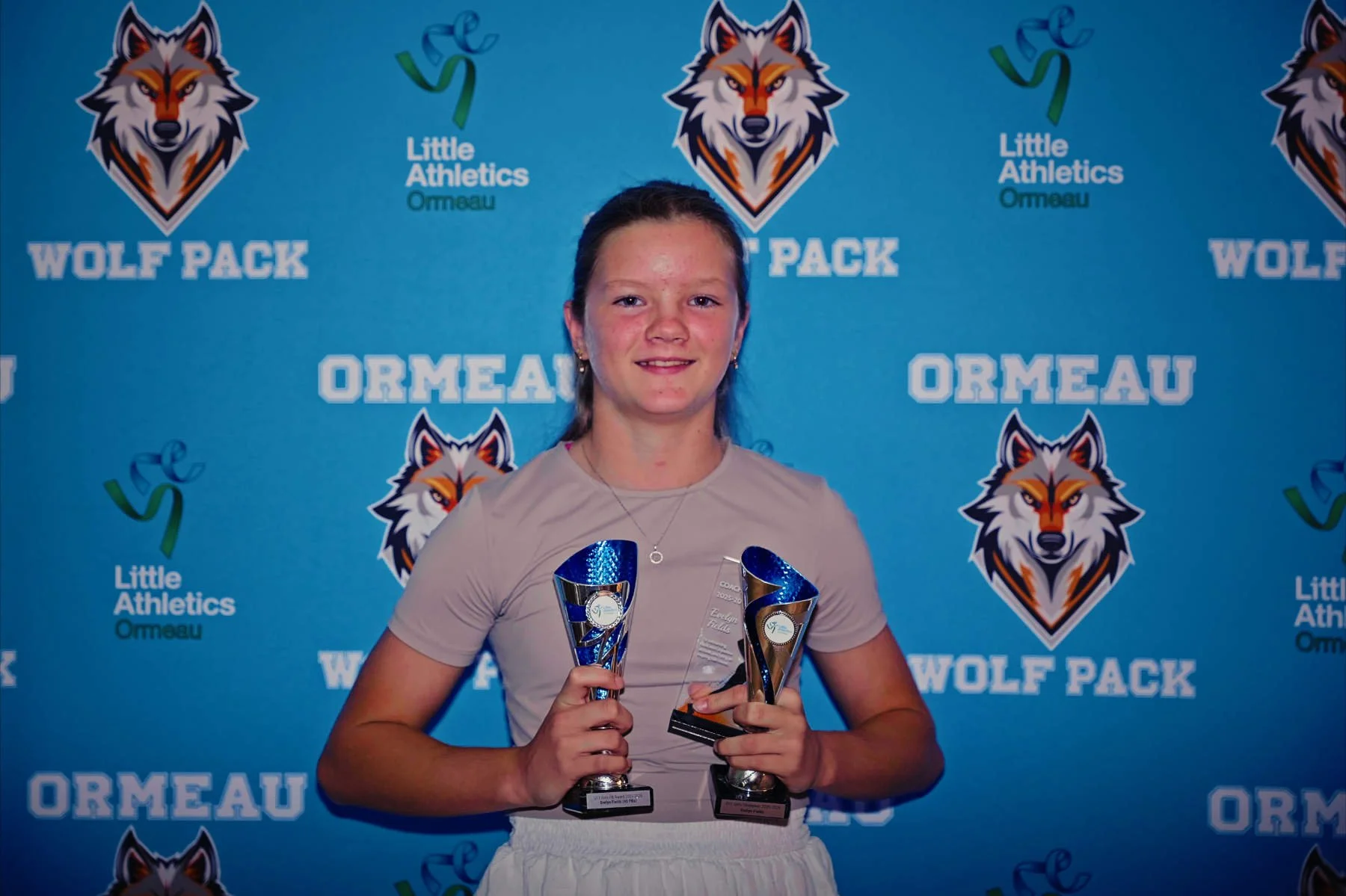 Youth girl with brown hair in a ponytail, wearing a beige athletic shirt, standing in front of a blue background with Wolf Pack, Little Athletics Ormeau logos, holding two trophies with one smiling expression.