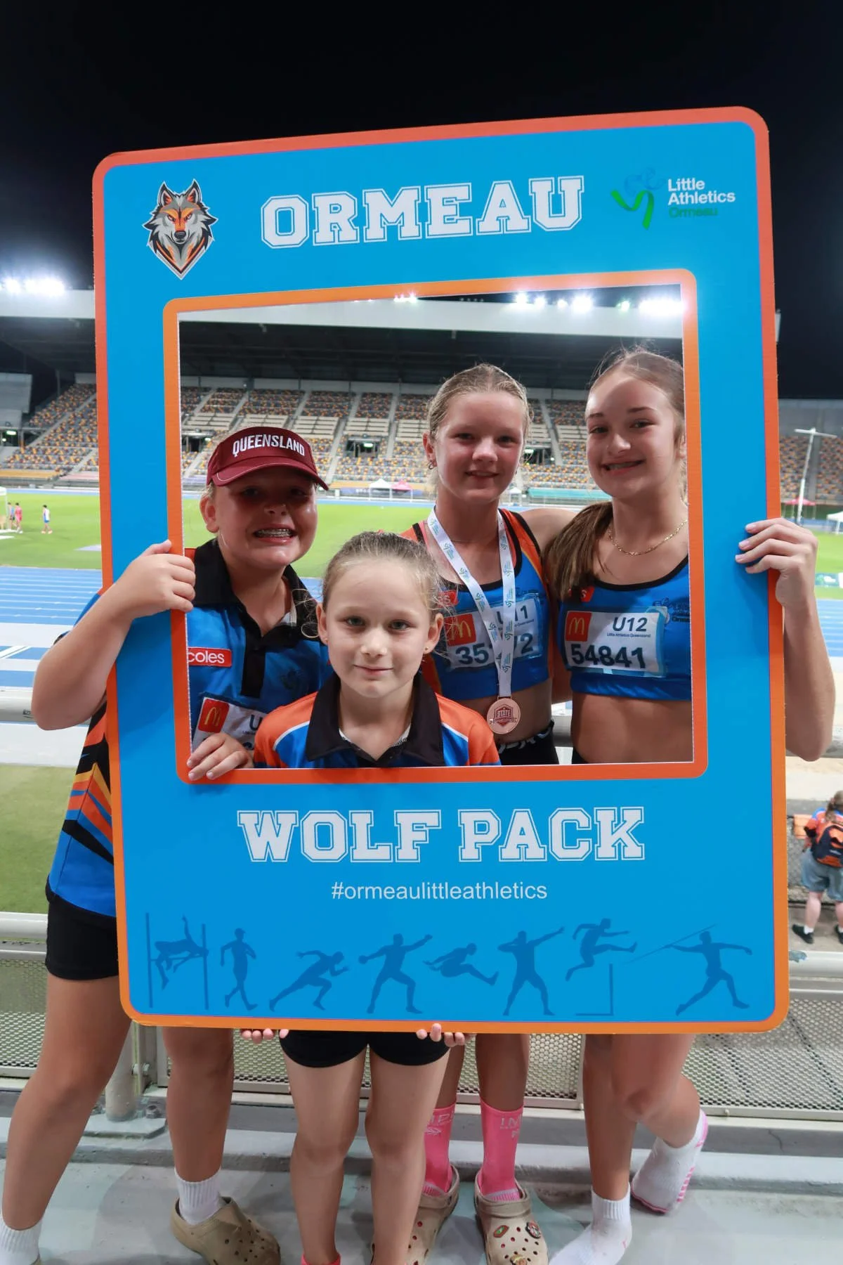 Four young girls at a sports event holding a blue frame with the text 'Ormeau Wolf Pack' and a hashtag '#ormeaultleathletics'. They are wearing athletic uniforms with various sponsor logos and medals. The background shows a stadium with a running track and field.