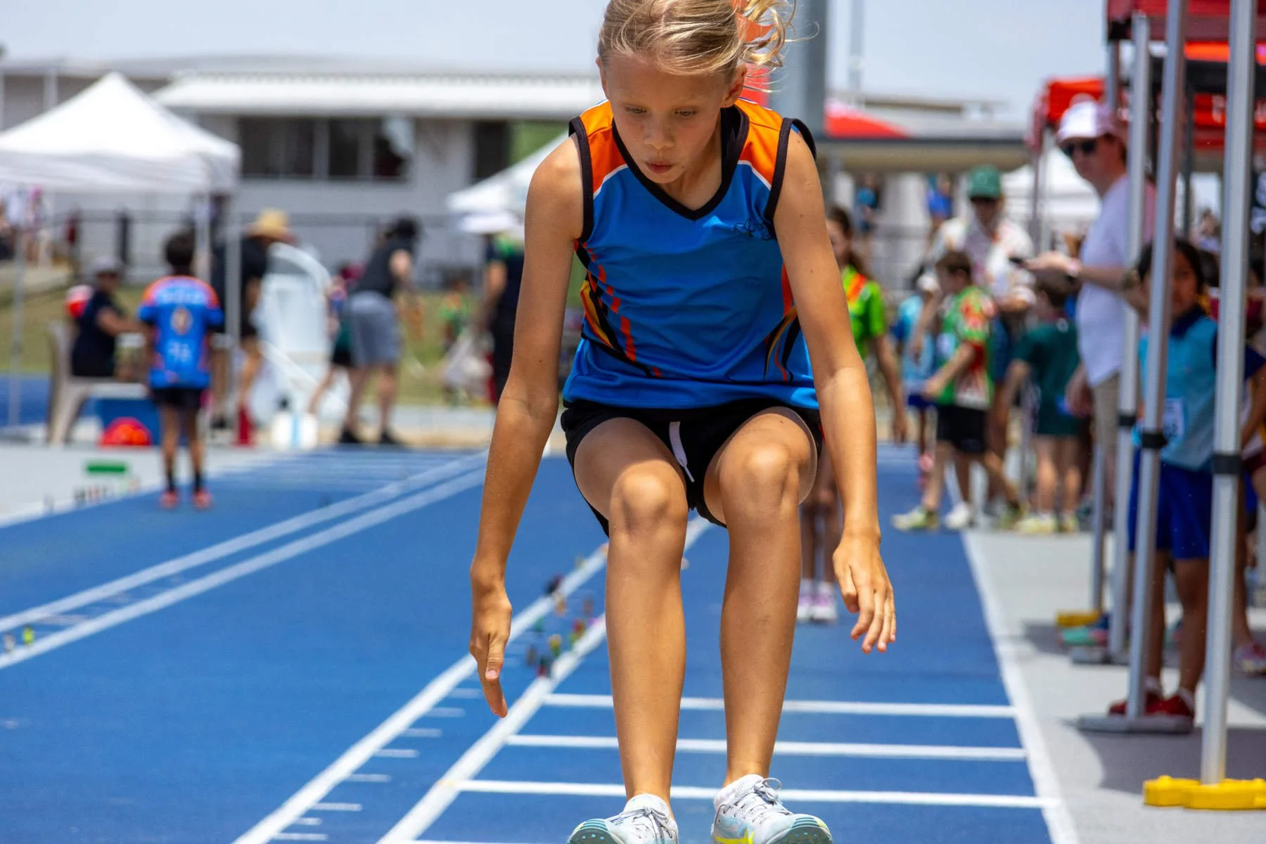 Young girl in athletic shorts and a blue, orange, and black sports top on a running track, crouched down, preparing to race at an outdoor track event, with spectators and tents in the background.