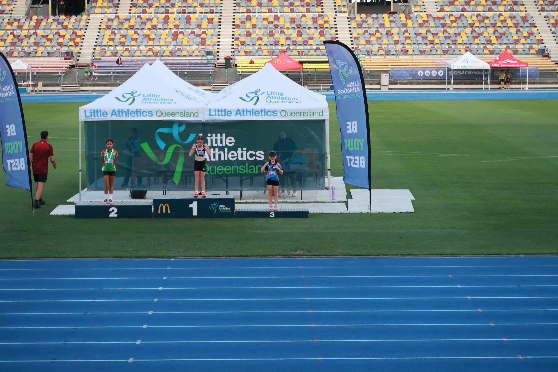 Children standing on winners' podium at a Little Athletics event in Queensland, Australia, with medals and sports attire, at an outdoor track stadium.