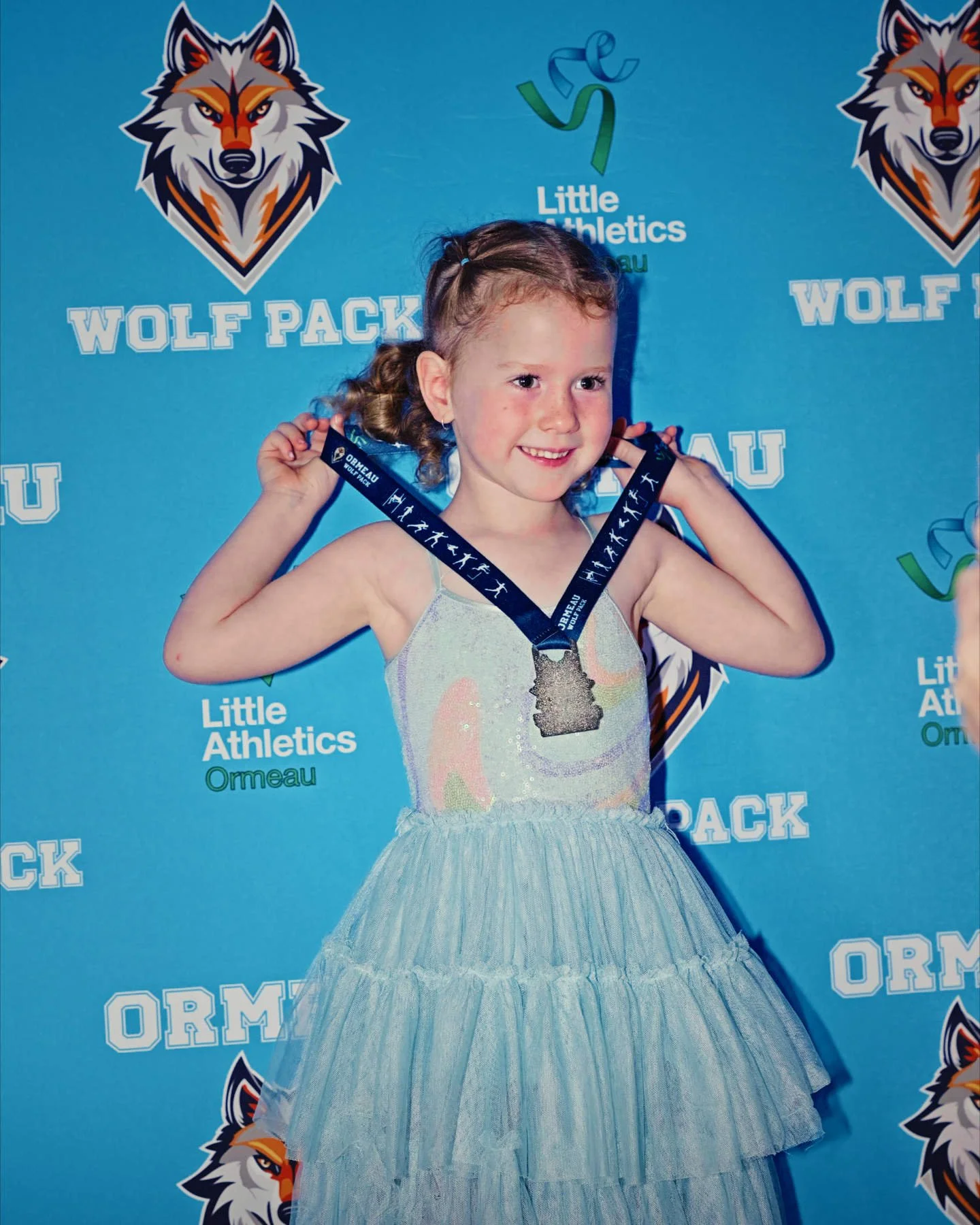 A young girl in a sparkly dress with a layered tulle skirt holding a medal at an athletic event, standing in front of a blue backdrop with logos for Wolf Pack, Little Athletics, and Ormeau.