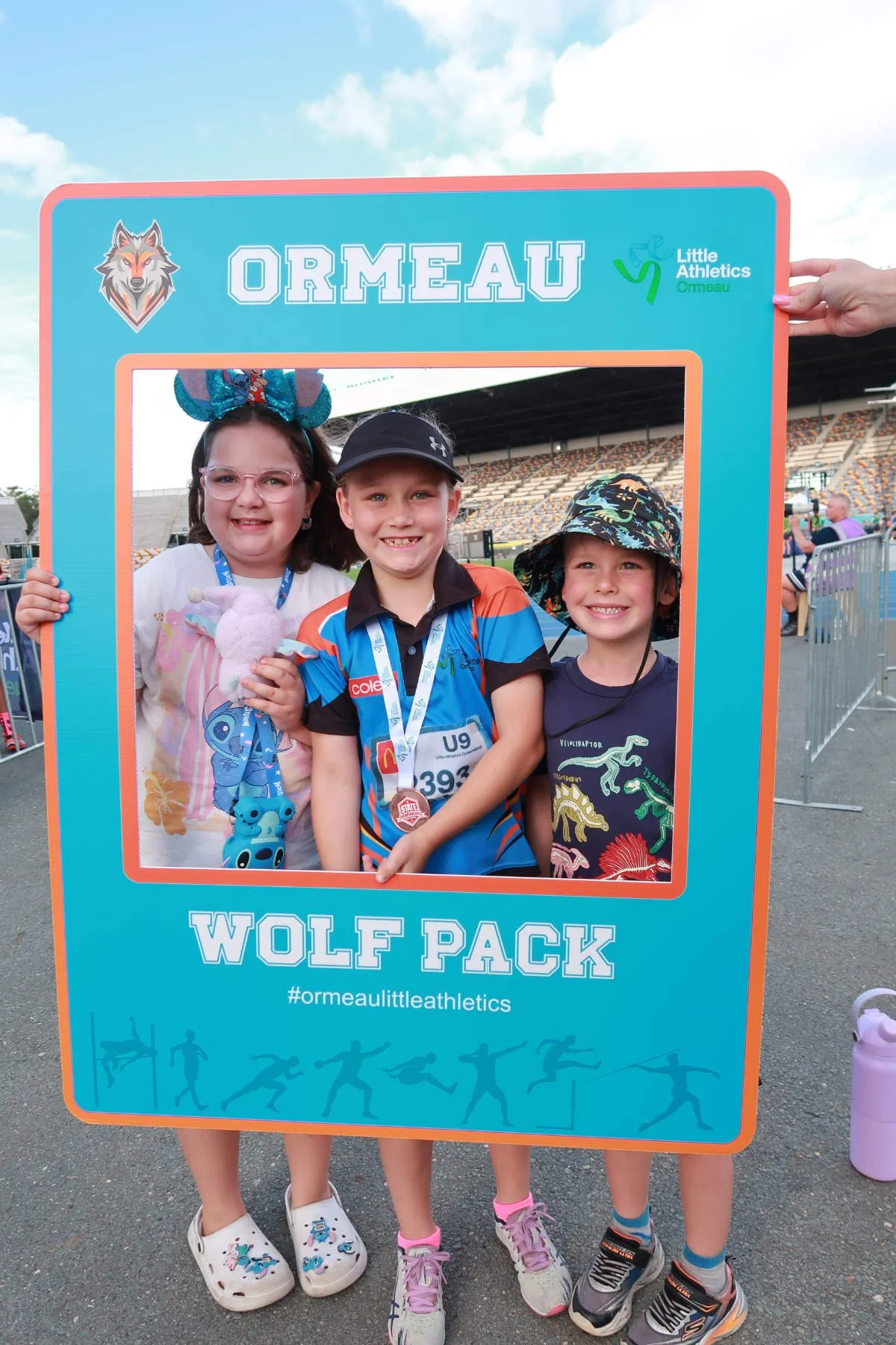 Three children standing together at an outdoor sports event, holding a large blue and orange frame with the words "Ormeau Wolf Pack" and hashtag #ormeaultleathletics. The children are smiling and wearing sports medals, and the background shows a stad