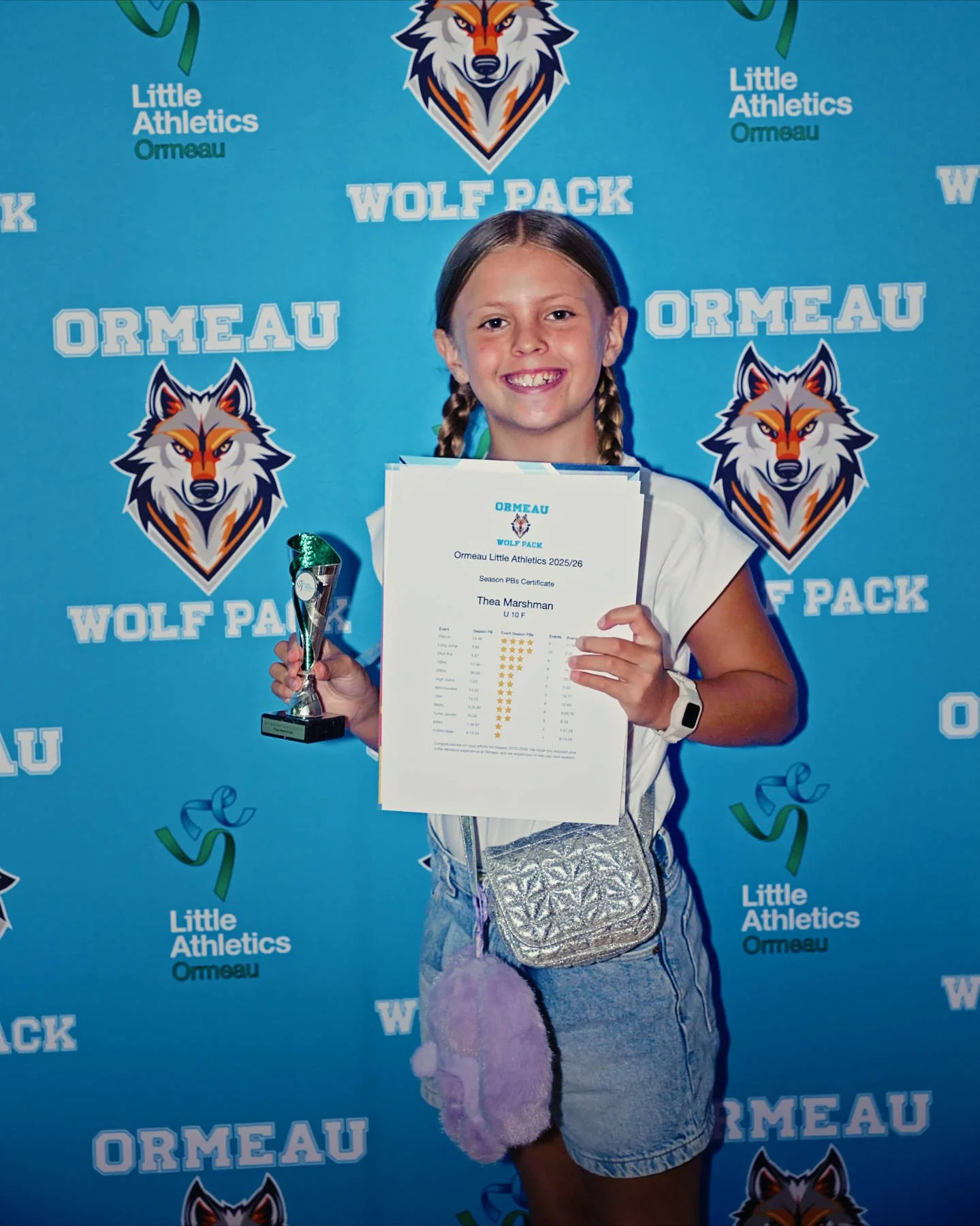 A young girl with braided hair smiling and holding a small trophy and a certificate, standing in front of a blue background with the words "Wolf Pack," "Ormeau," and "Little Athletics," along with wolf logos.