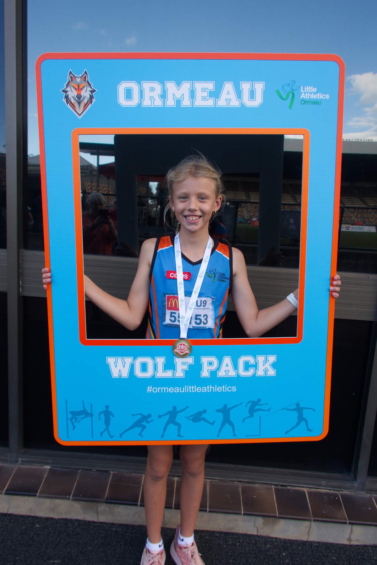 Young girl at a track meet holding a large foam frame with the words 'Wolf Pack' and hashtags for Little Athletics Ormeau. She is wearing a blue sports jersey with a medal around her neck, smiling at the camera.