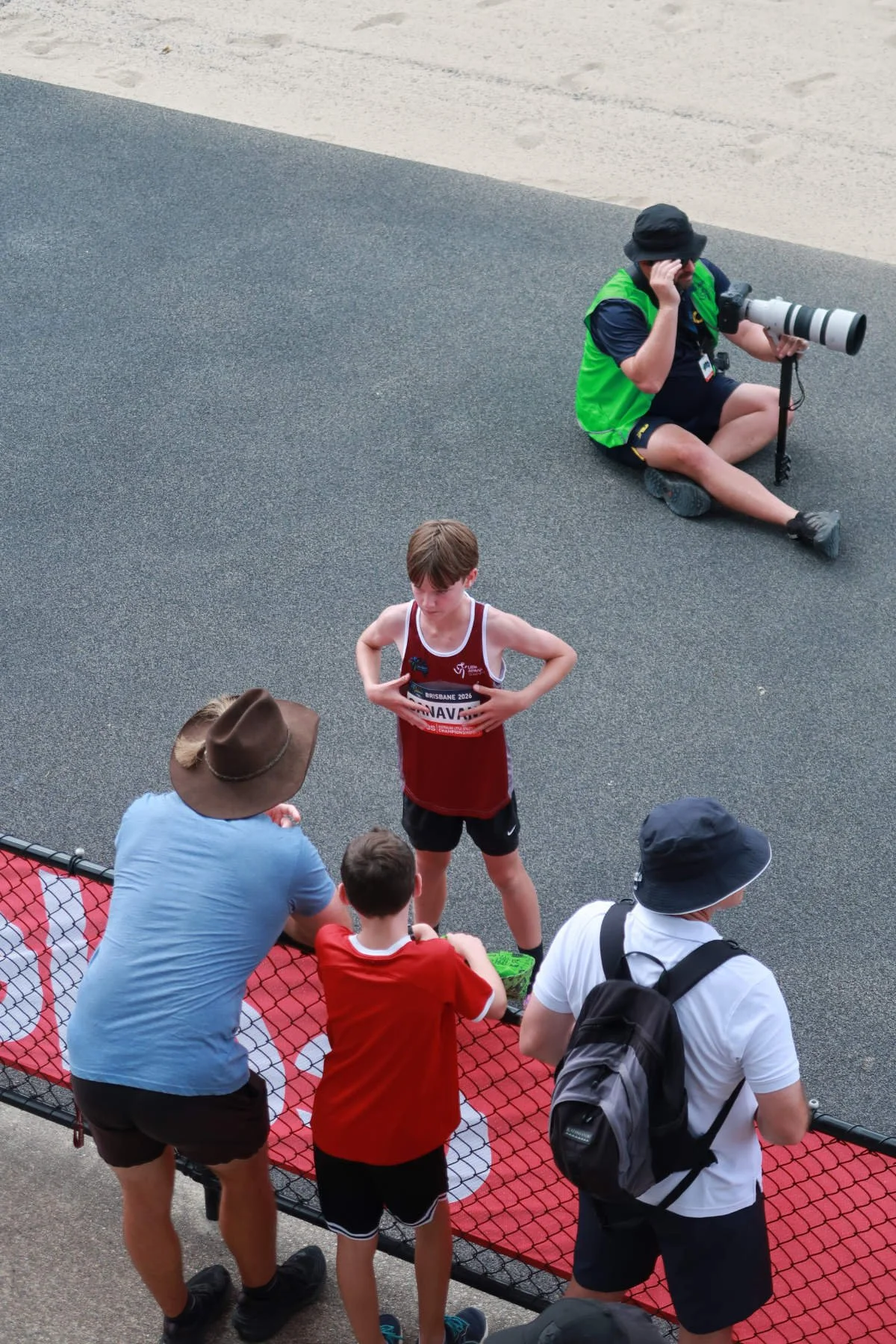 A young male athlete with a red tank top and black shorts standing on a track, surrounded by spectators, while a photographer with a large camera lens sits on the ground nearby, taking photos.