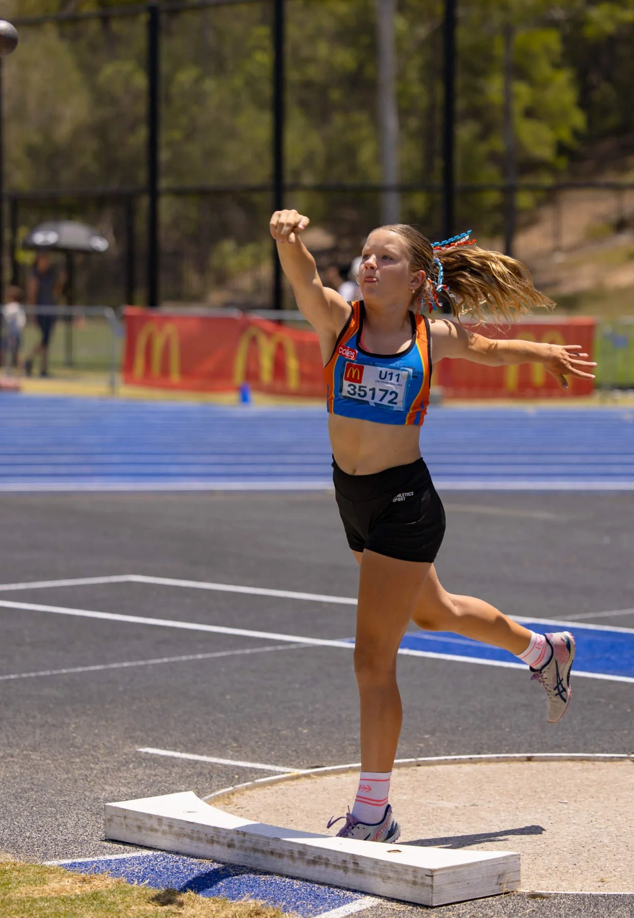 A young female athlete in a blue and orange sports uniform running on a track during a race, with a numbered bib, using a long jump sandpit.