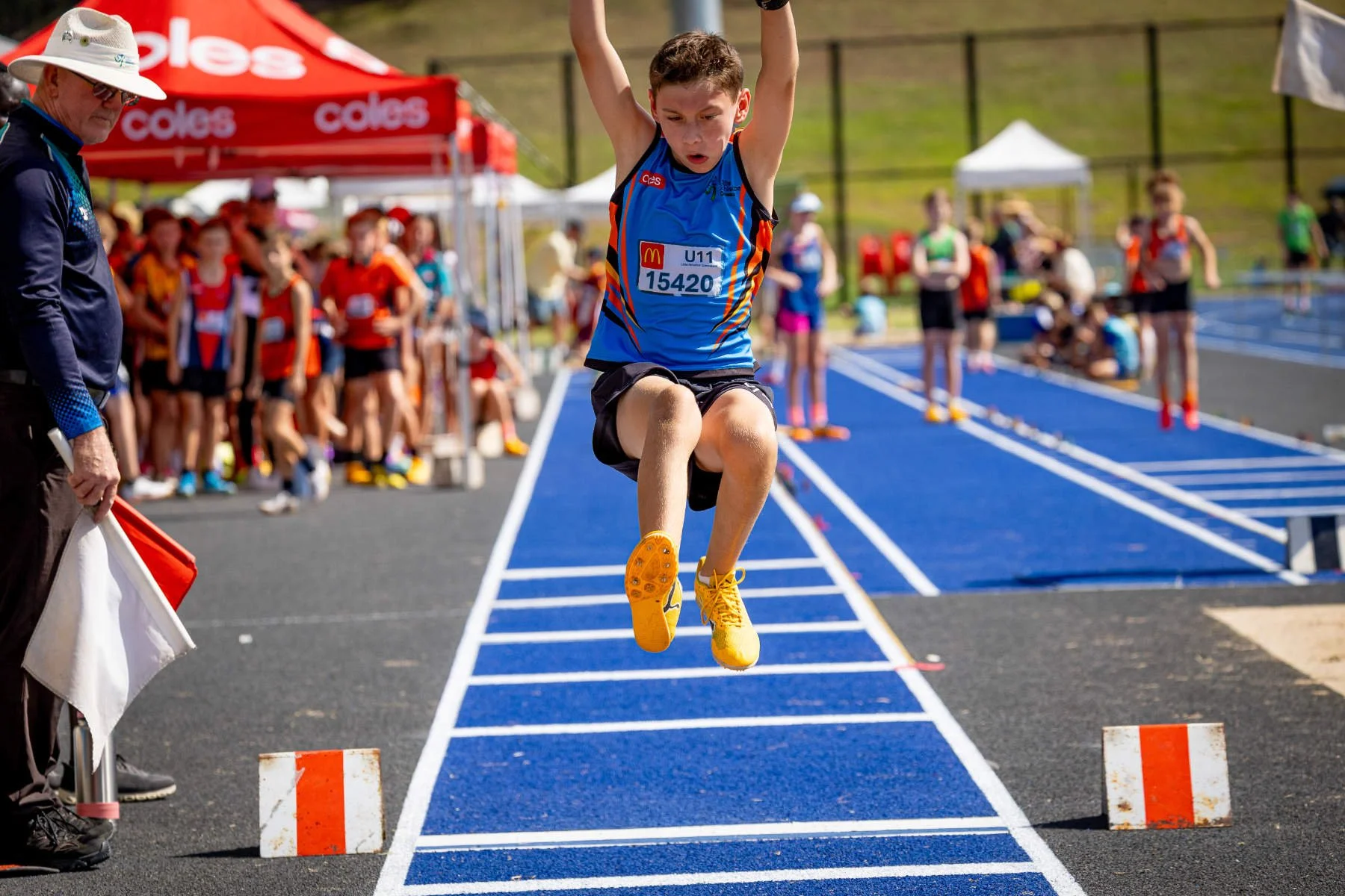 Young boy in a blue and black sports uniform with a race bib number 15420 jumping during a track and field competition at a blue running track, with spectators and athletes in the background.