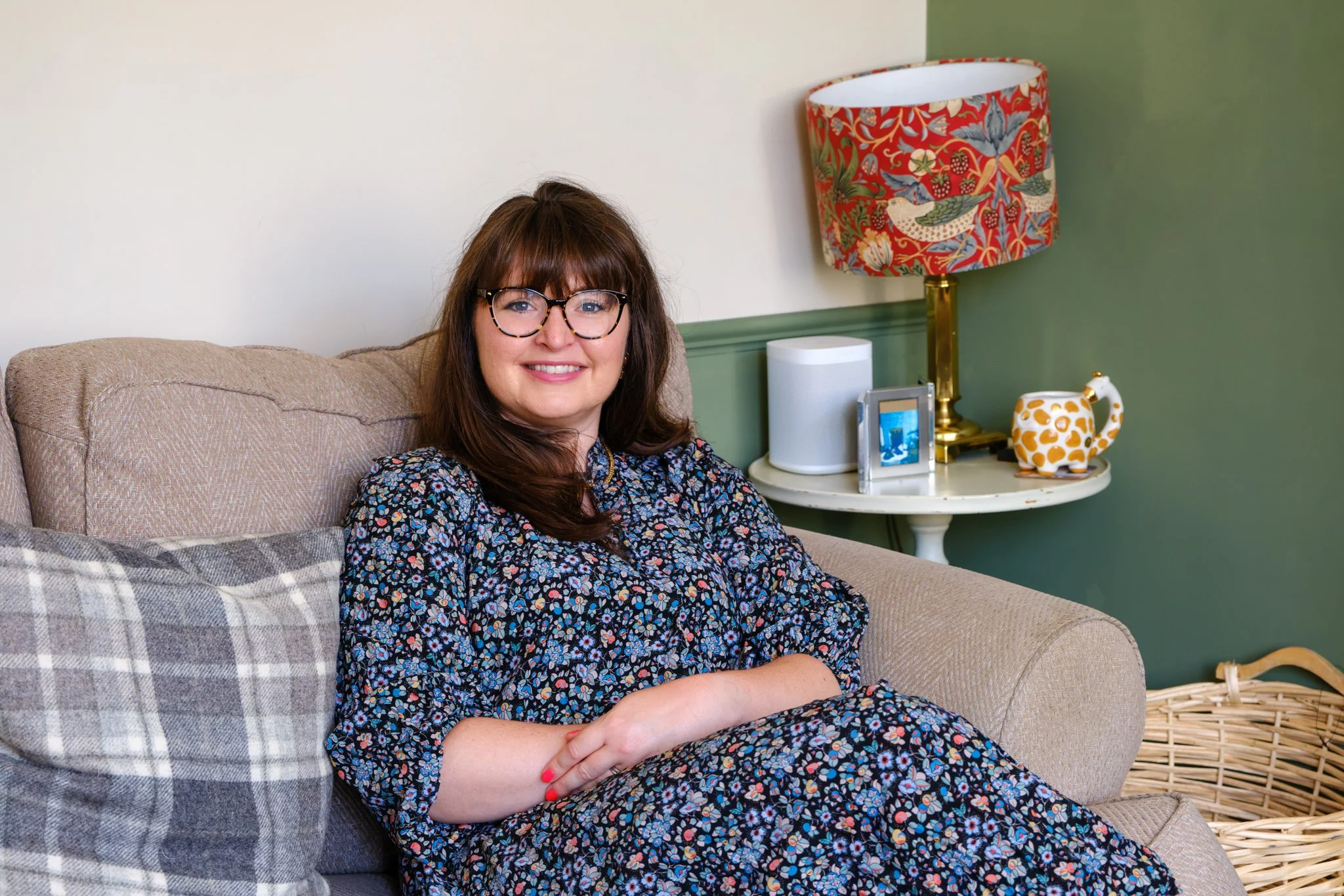 A woman with glasses and brown hair sitting on a beige sofa in a living room, smiling at the camera. There is a green wall, a colorful lampshade, a white speaker, a framed photo, a teapot, and a wicker basket on a side table.