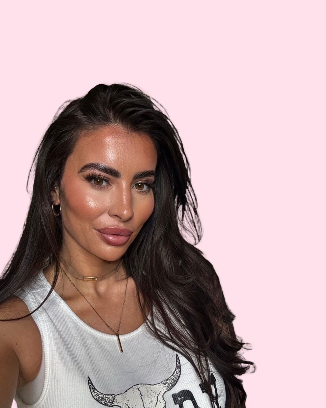A woman with long dark hair, wearing makeup, a white tank top with a bull skull graphic, and layered necklaces, posing against a light pink background.