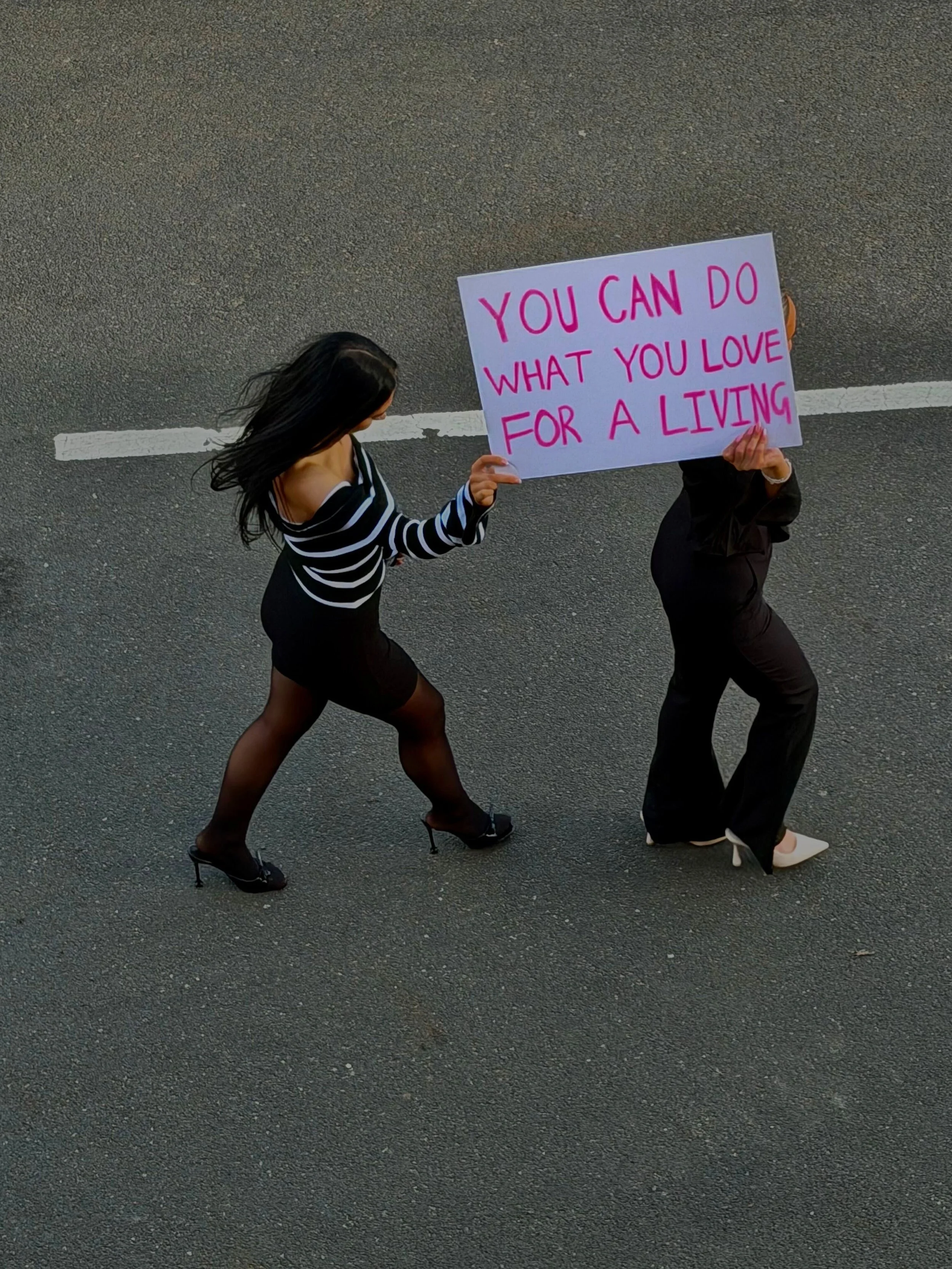 A woman wearing black high heels, black stockings, and a striped black and white top walking on a street, holding the hand of another person who is holding a bright pink sign. The sign reads 'You can do what you love for a living.'