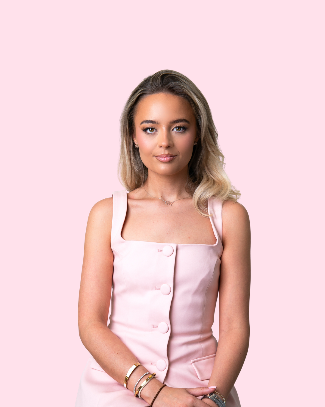 A young woman with blonde wavy hair and light makeup, wearing a sleeveless pink dress with large buttons, standing against a light pink background.