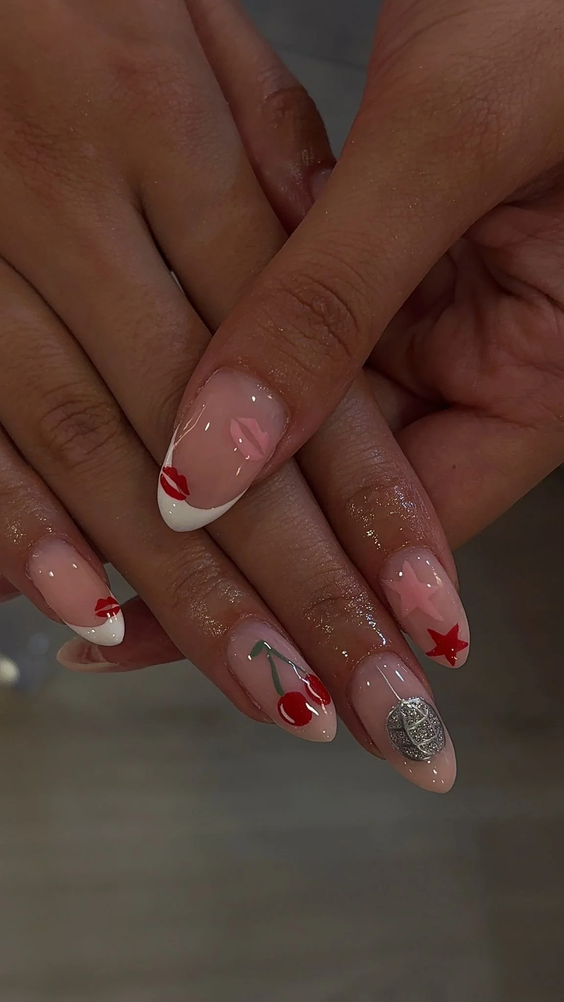 Close-up of hands with decorated nails featuring Christmas-themed designs, including cherries, stars, lips, and a silver pattern.