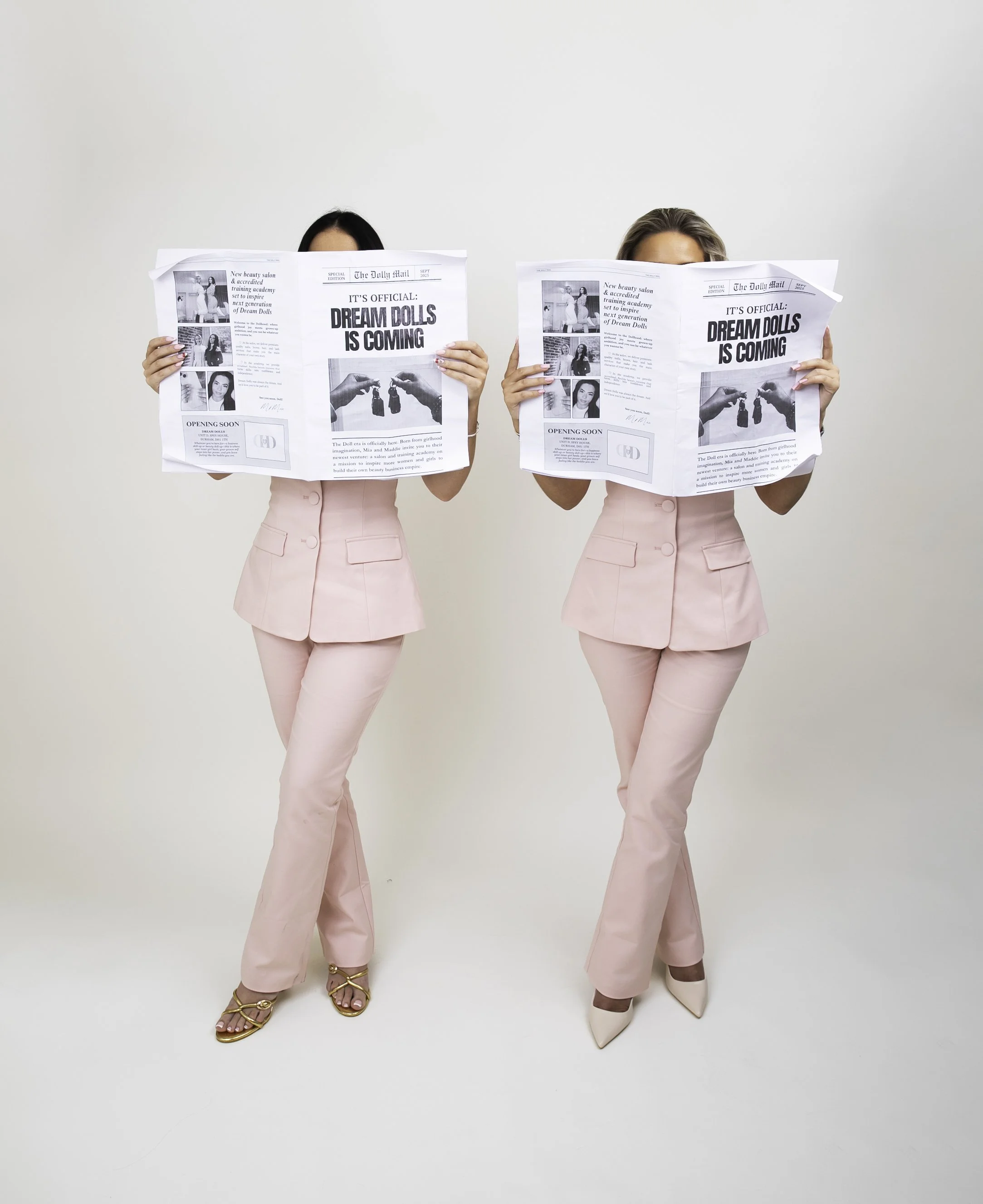 Two women dressed in pink pantsuits holding newspapers covering their faces against a plain white background.