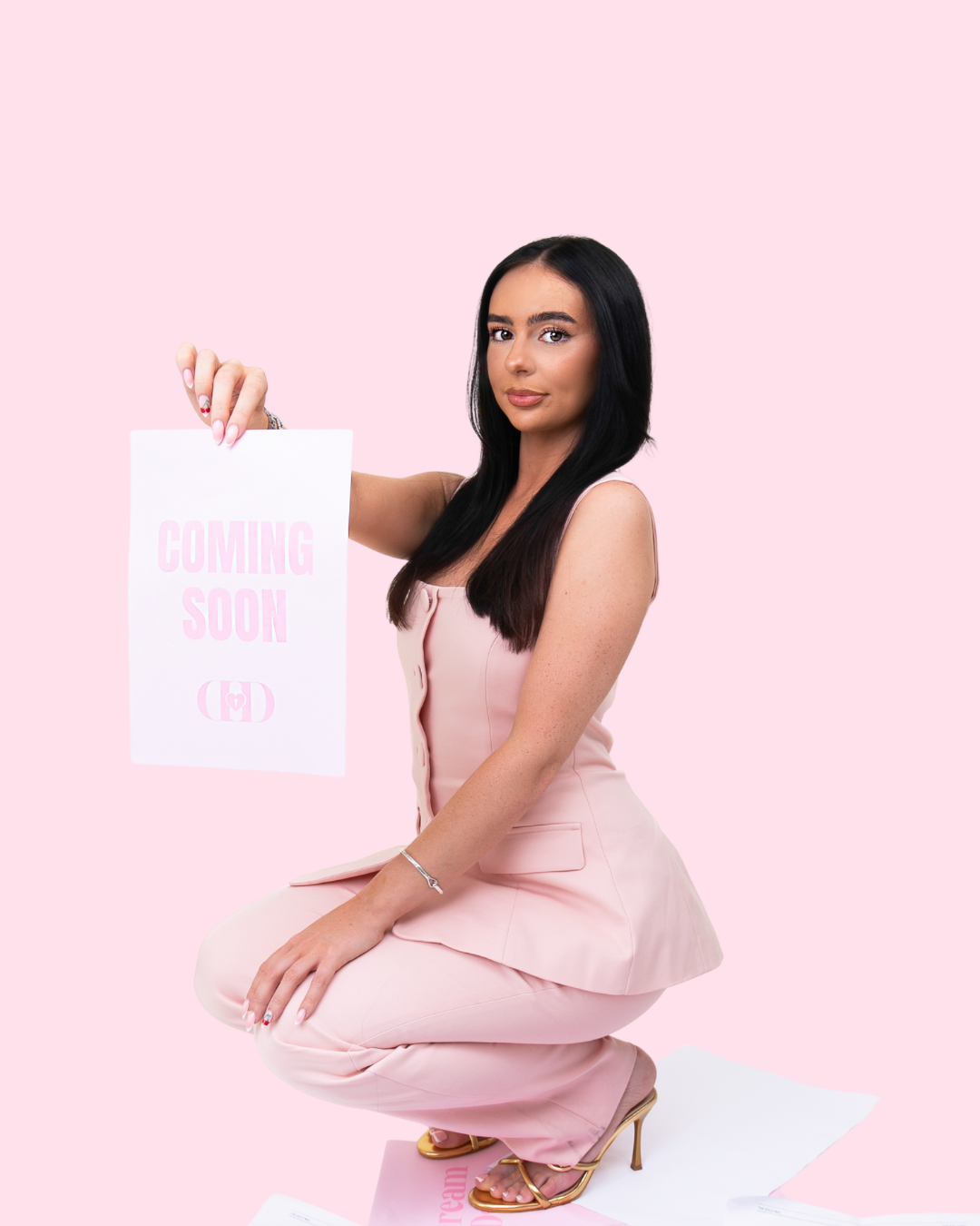 A woman in a pink outfit kneeling on a pink background, holding a sign that says 'Coming Soon' with pink lettering.