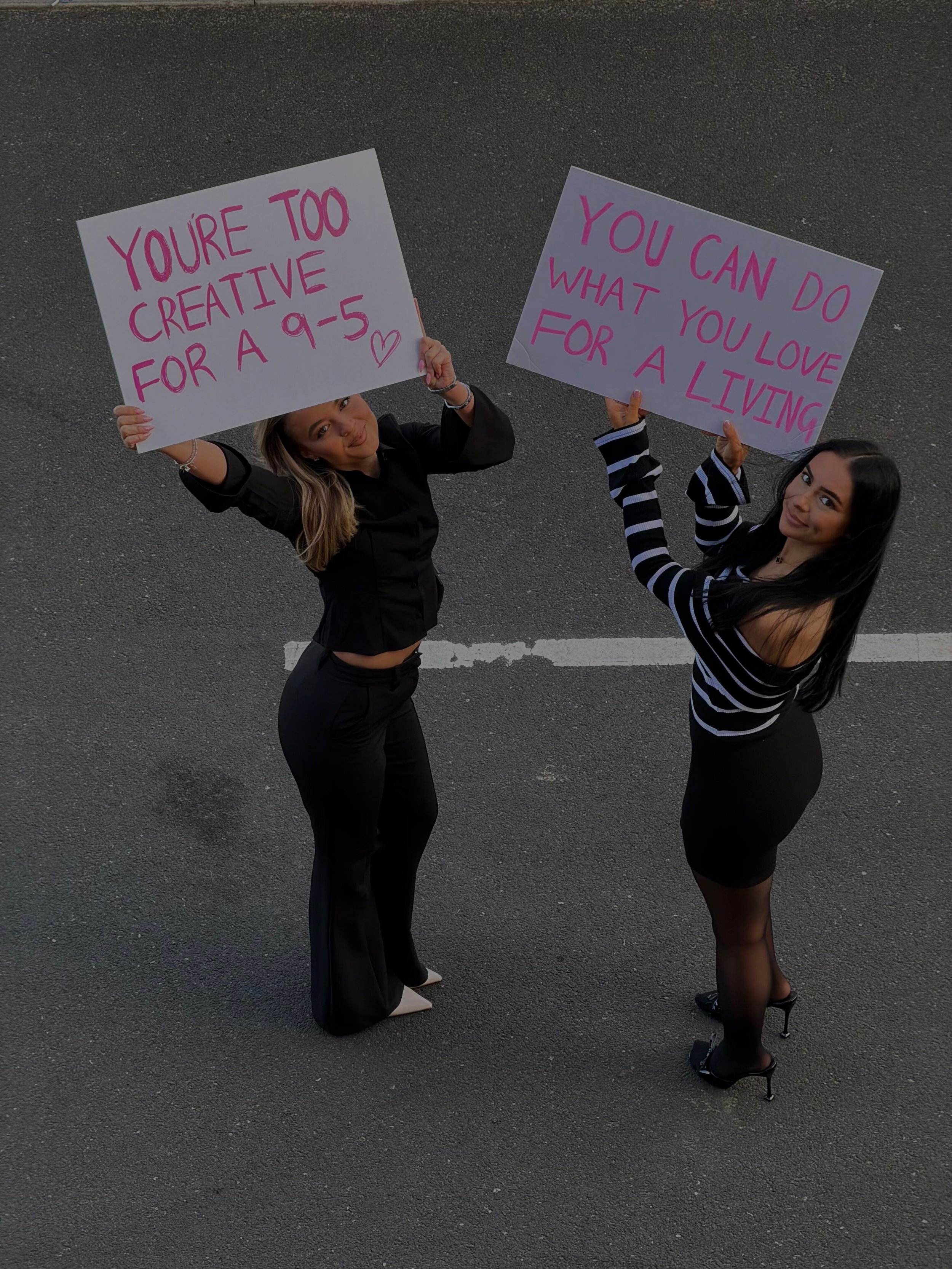 Two women standing on an asphalt road, holding large pink signboards with motivational messages. The woman on the left has blonde hair, dressed in black, and the woman on the right has black hair, dressed in a black and white striped top, black skirt, and high heels.