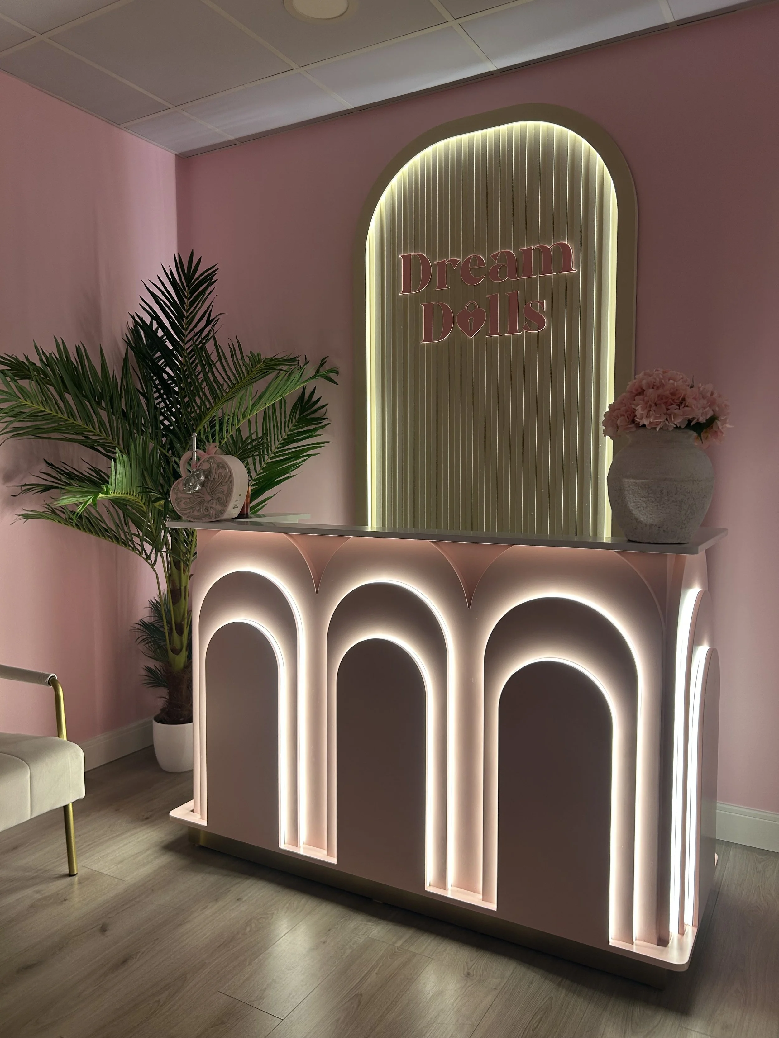 Decorative pink wall with illuminated sign reading 'Dream Dolls', pink flowers in a white vase on a table, a large green plant, and a white chair with gold legs in a modern interior.