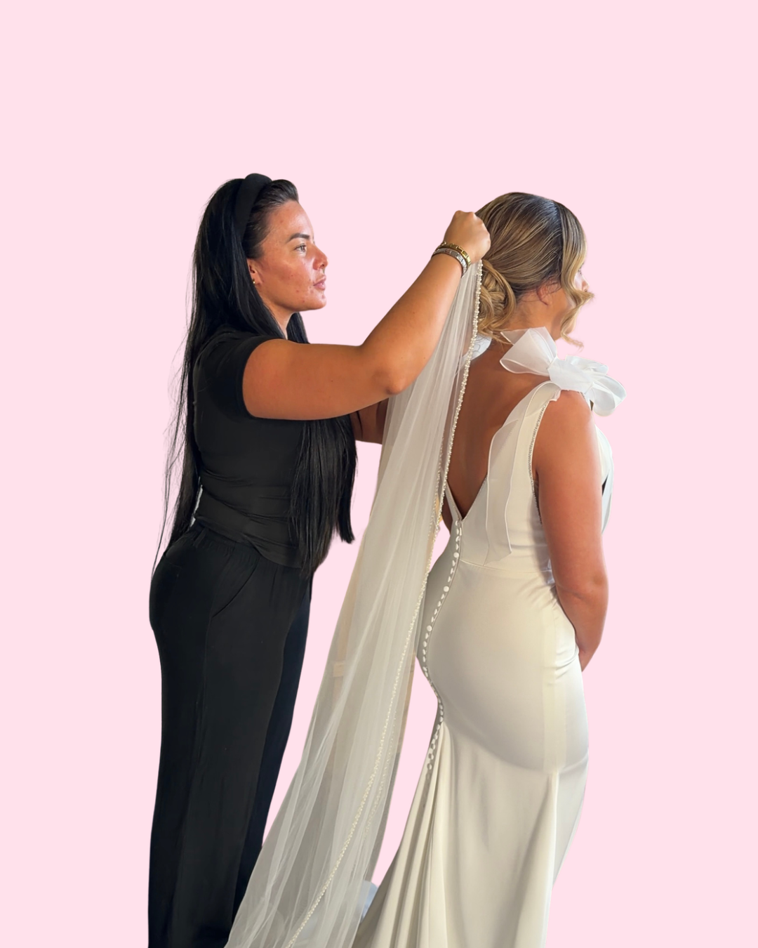 A woman in a white wedding dress is being assisted with her veil by a woman dressed in black, against a pink background.