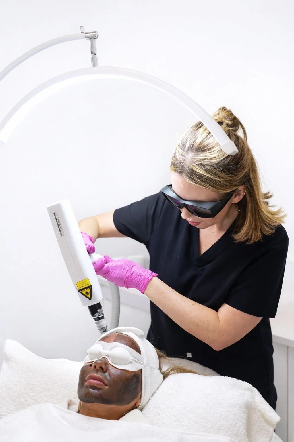 A woman receiving a facial treatment with a laser device while wearing sunglasses and gloves, lying on a treatment bed with a facial mask applied.