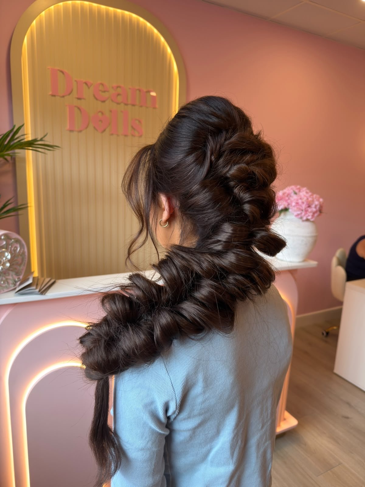 A woman with long dark hair styled in voluminous curls and a partial braid, viewed from behind, in a pastel pink and yellow-lit salon with a sign that reads 'Dream D(ills)' in pink letters. There is a white table with pink flowers in a white vase and a person in a black top sitting in the background.