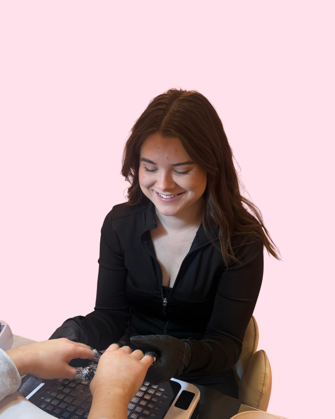 A woman with brown hair smiling as she gets a manicure from a technician wearing black gloves at a salon.