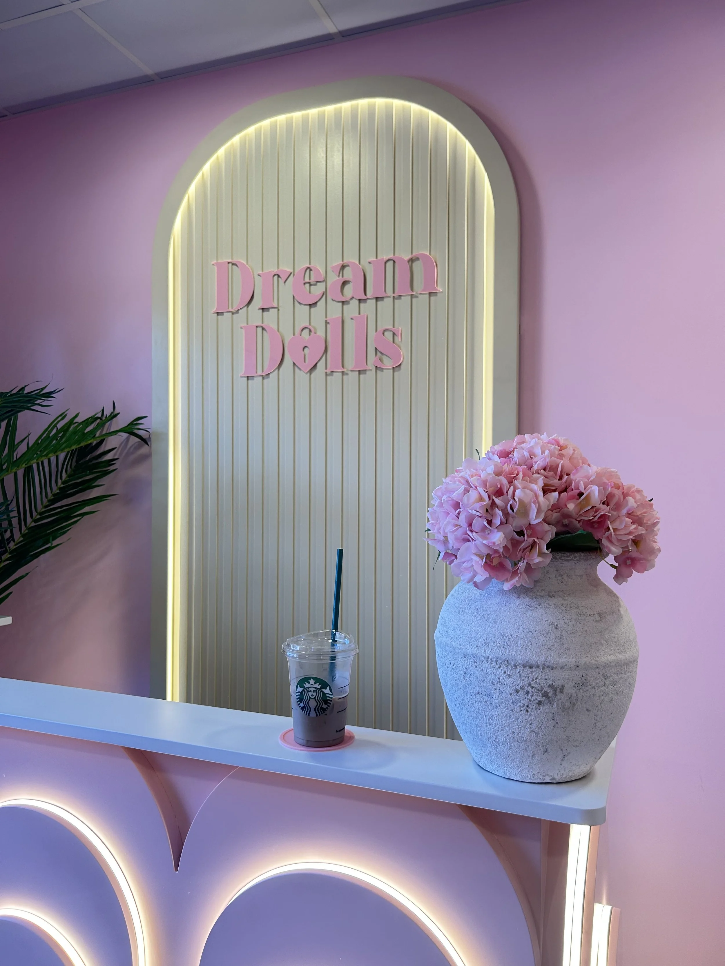 A pastel pink and purple decor area featuring a sign that says 'Dream Dolls' in pink letters. There is a large white textured vase with pink hydrangea flowers, a Starbucks cup with a straw, and a tropical plant on a counter.