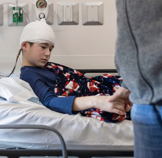 A young boy with a white knit hat in a hospital bed, wearing a blue shirt, covered with a colorful blanket, being held by a person standing nearby.