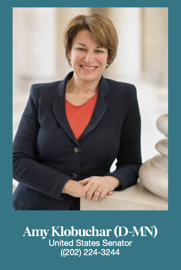 Photograph of Amy Klobuchar, a middle-aged woman with short brown hair, wearing a dark blazer over a red top, smiling and standing indoors near a white sculpture.