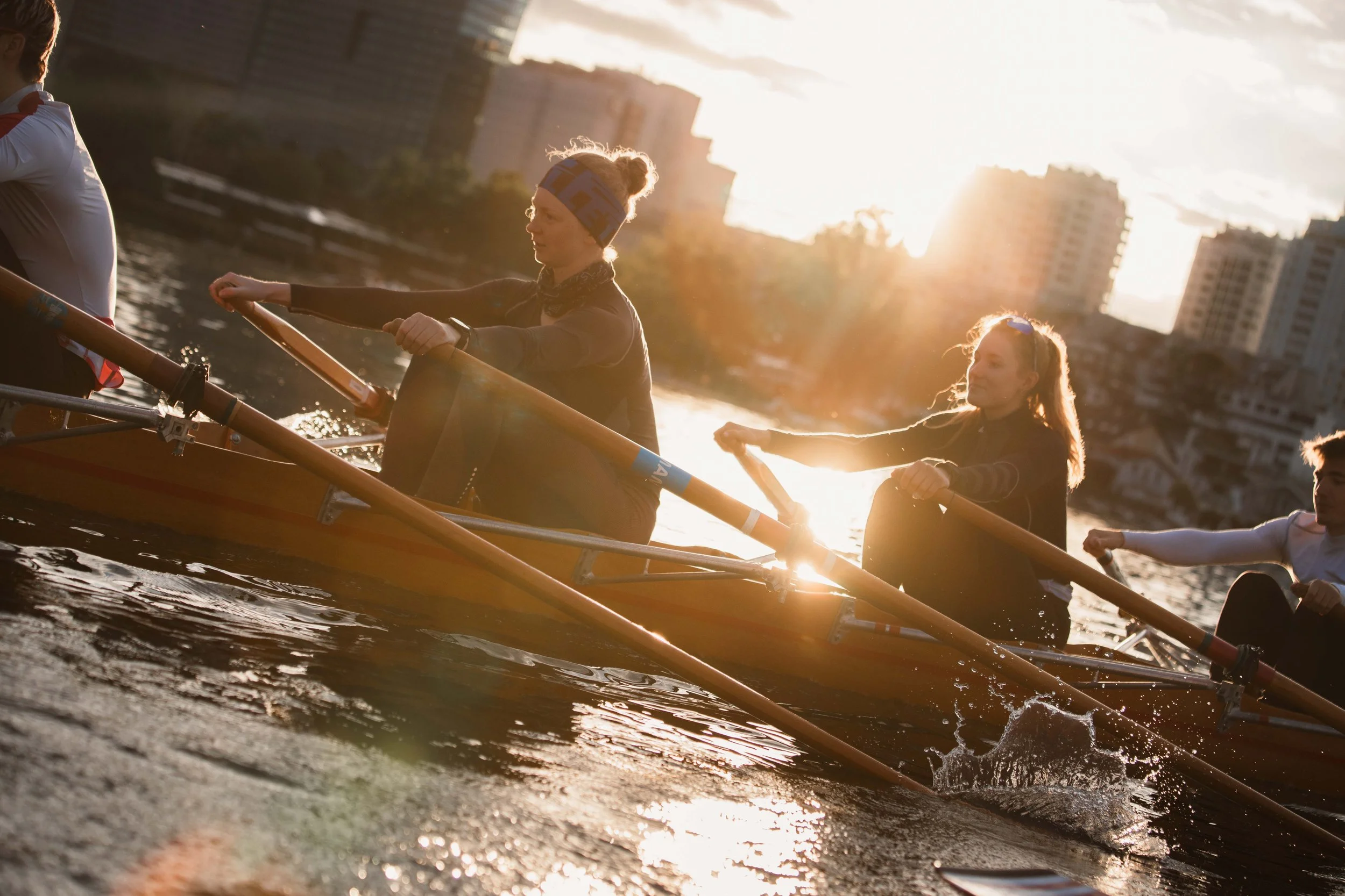 Menschen beim Rudern auf der alten Donau bei Sonnenuntergang, mit Hochhäusern im Hintergrund.