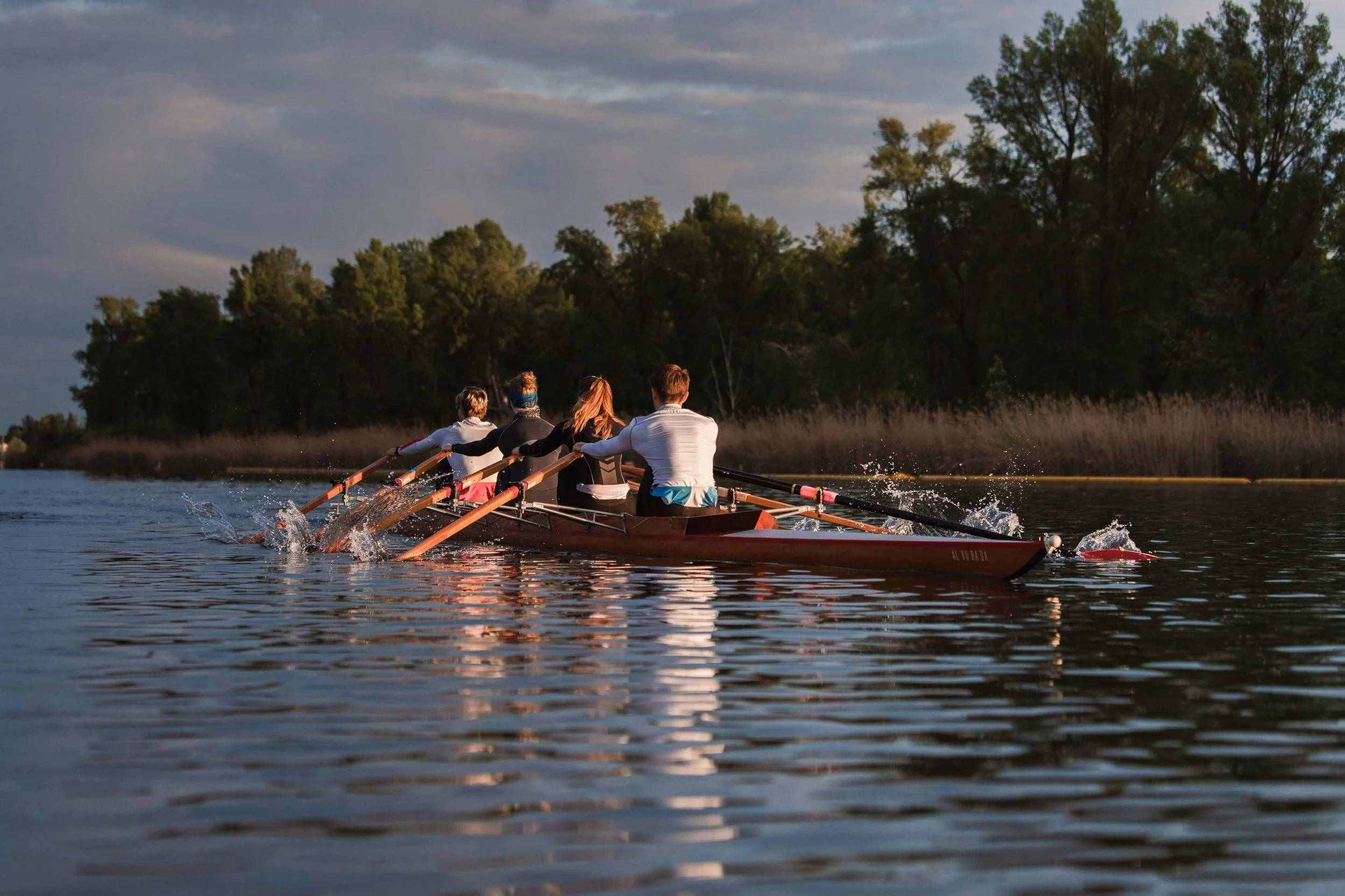 Eine Gruppe von vier Menschen beim Rudern auf einem Fluss, umgeben von Bäumen und bewölktem Himmel.