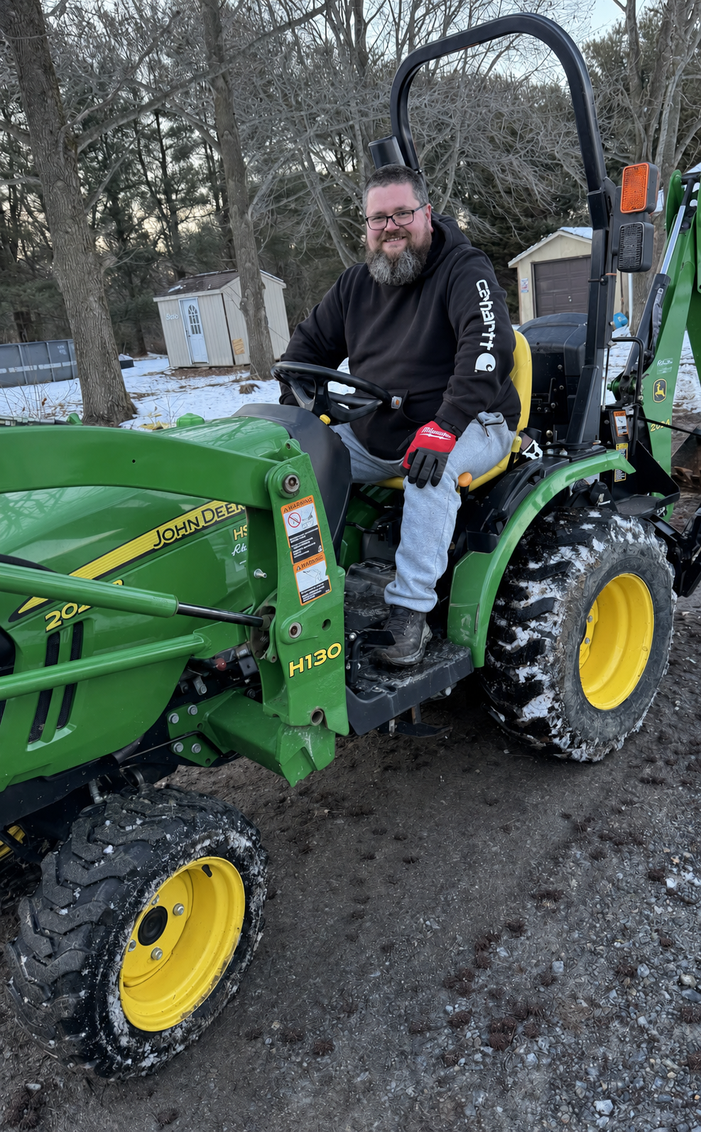 A smiling man with glasses and a beard sitting on a green John Deere tractor in a snowy outdoor setting, wearing a black hoodie, gray sweatpants, and red gloves.