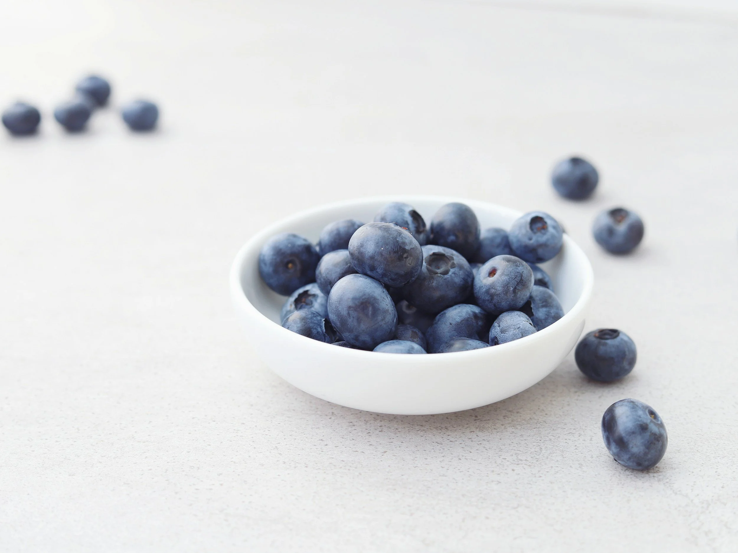 Close-up of fresh blueberries in a white bowl on a white surface, with some blueberries scattered around.