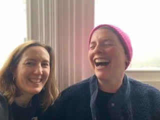 Emily and Gabrielle  smiling and laughing together indoors,  Gabrielle is wearing a pink beanie.