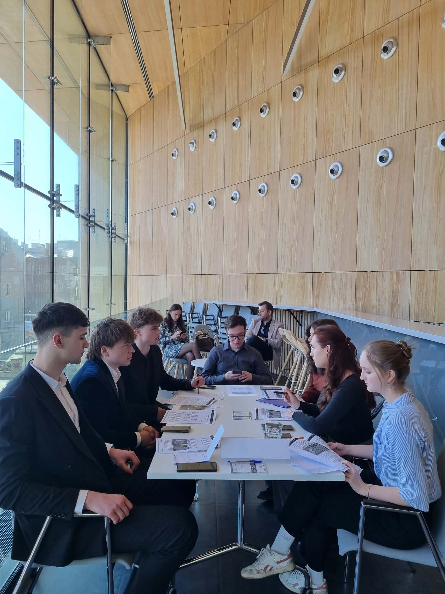 A group of people seated around a rectangular table in a modern, bright room with large windows. They have documents, electronic devices, and cups on the table and appear to be engaged in a meeting or discussion.