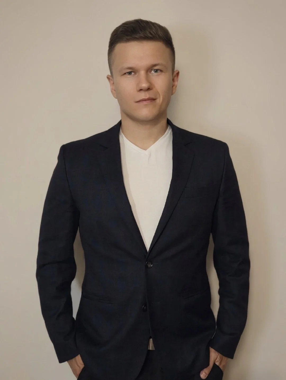 A young man with short brown hair, wearing a dark suit jacket over a white shirt, standing against a beige wall.