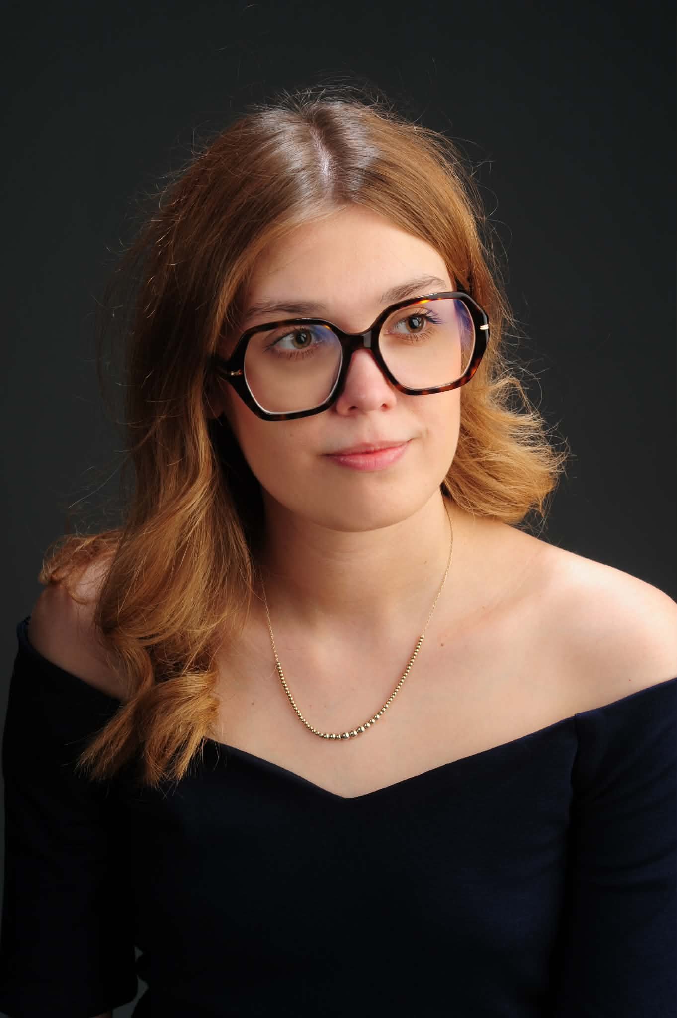 Close-up portrait of a young woman with red hair, wearing large tortoiseshell glasses, a black off-the-shoulder top, a gold necklace with small beads, and a subtle smile, against a dark background.