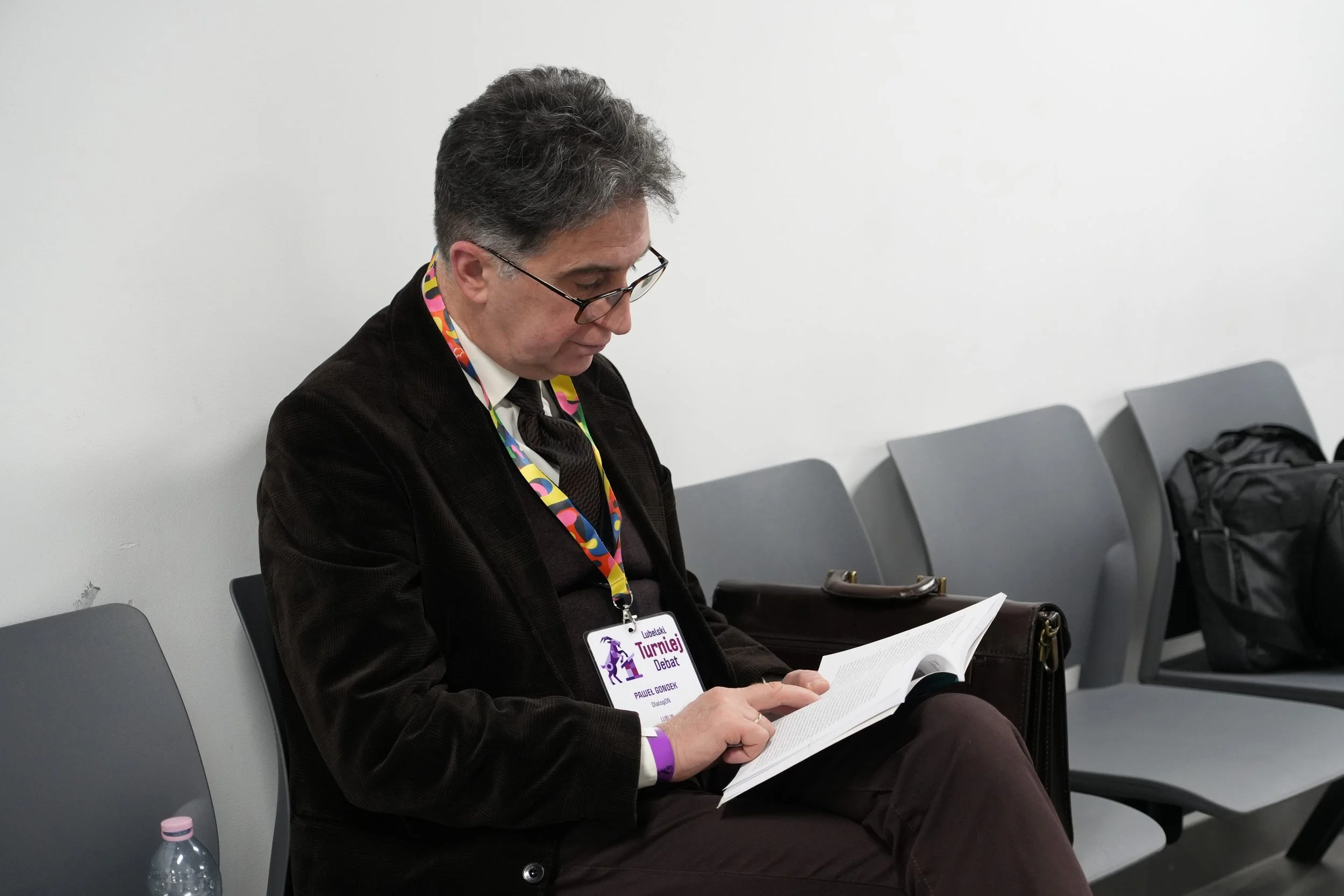 A man with dark gray hair and glasses wearing a black jacket and a colorful lanyard, sitting in a row of gray chairs against a white wall, reading a document with a water bottle on the floor beside him.