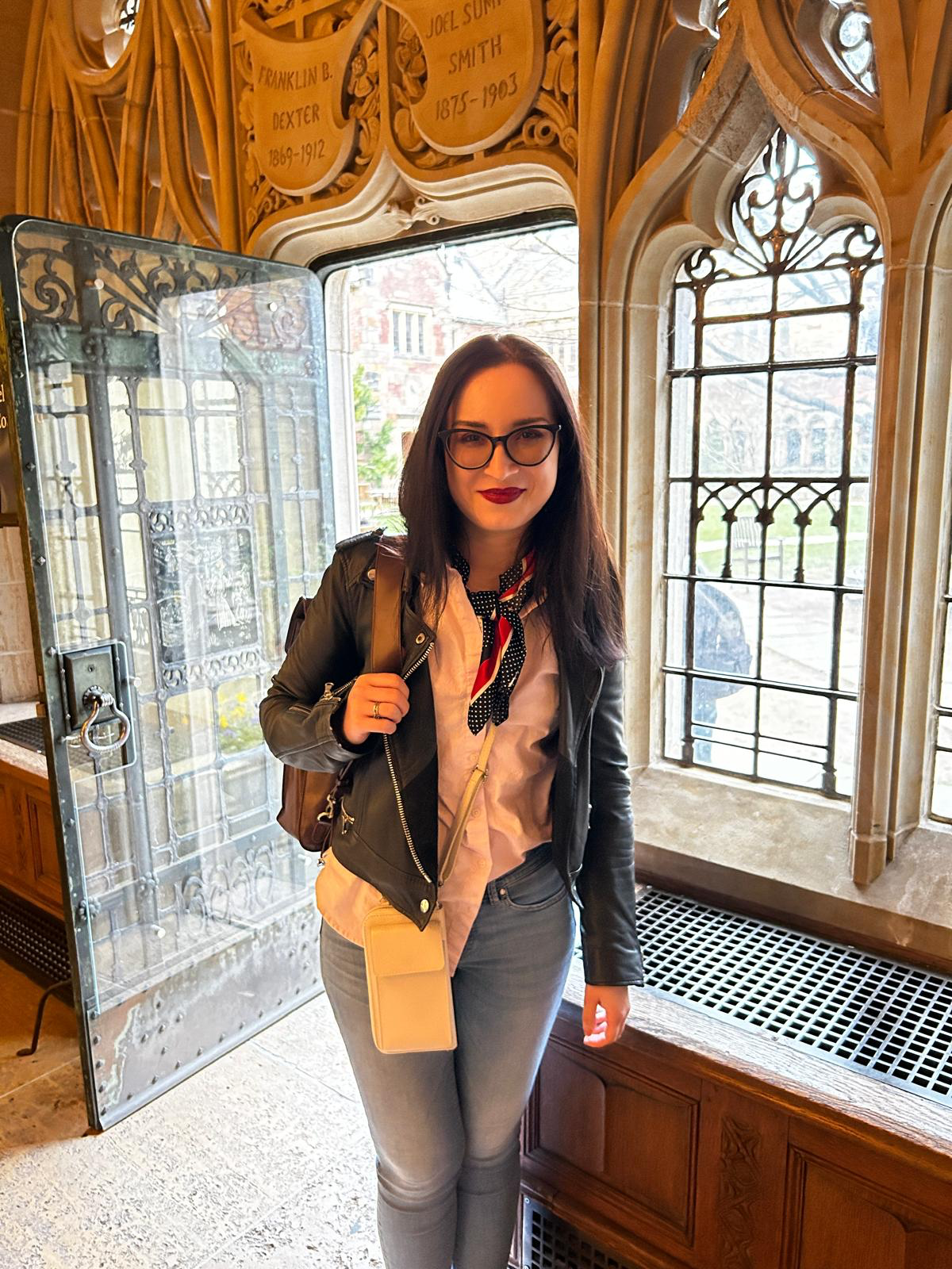 A woman with black hair and glasses standing indoors near a window, wearing a black leather jacket, a white blouse, gray jeans, and a black-and-white polka dot scarf, carrying a beige crossbody bag and a backpack.