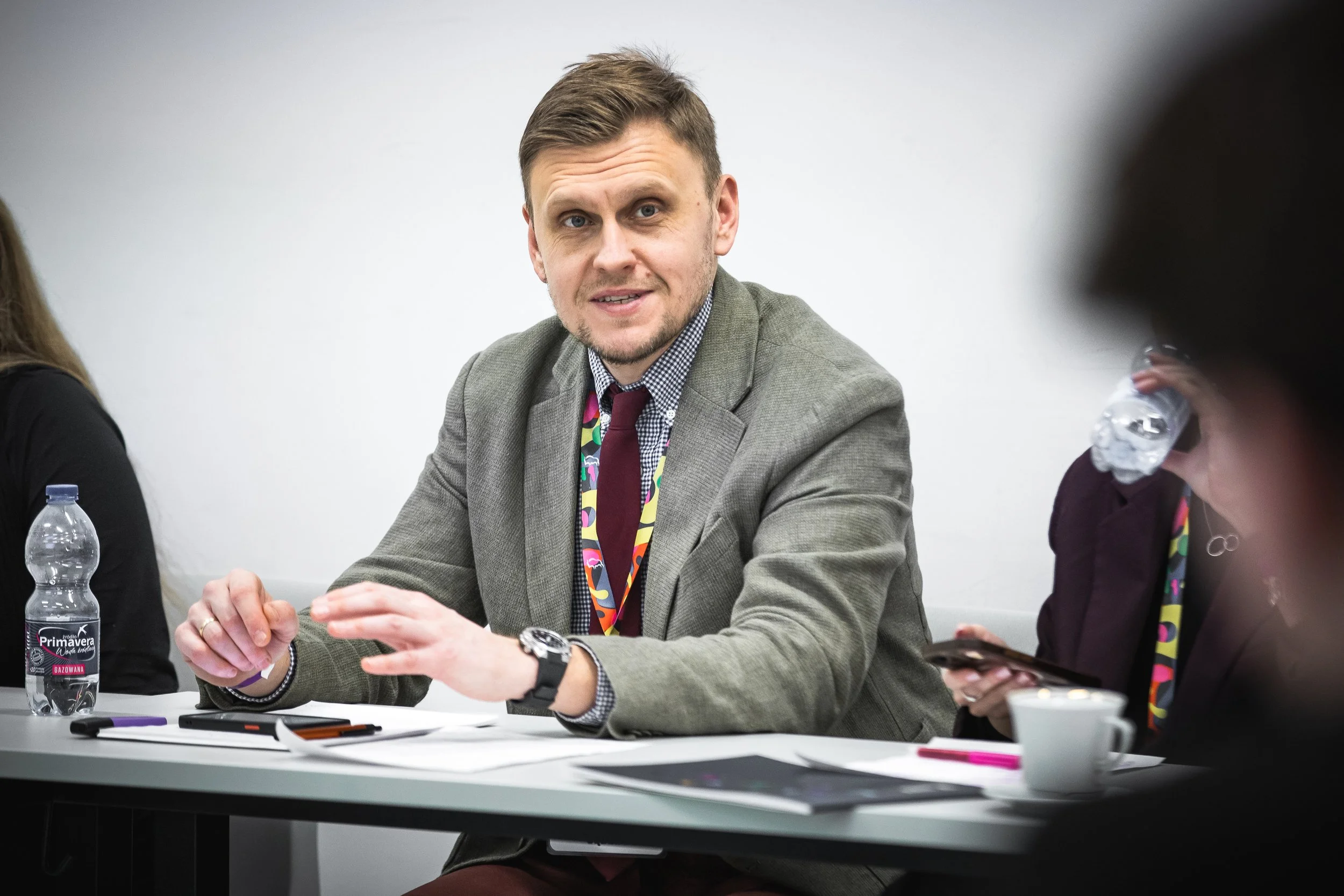 A man in a gray blazer and checkered shirt sitting at a table with other people, gesturing with his hands during a discussion. Bottled water, papers, and writing utensils are on the table.