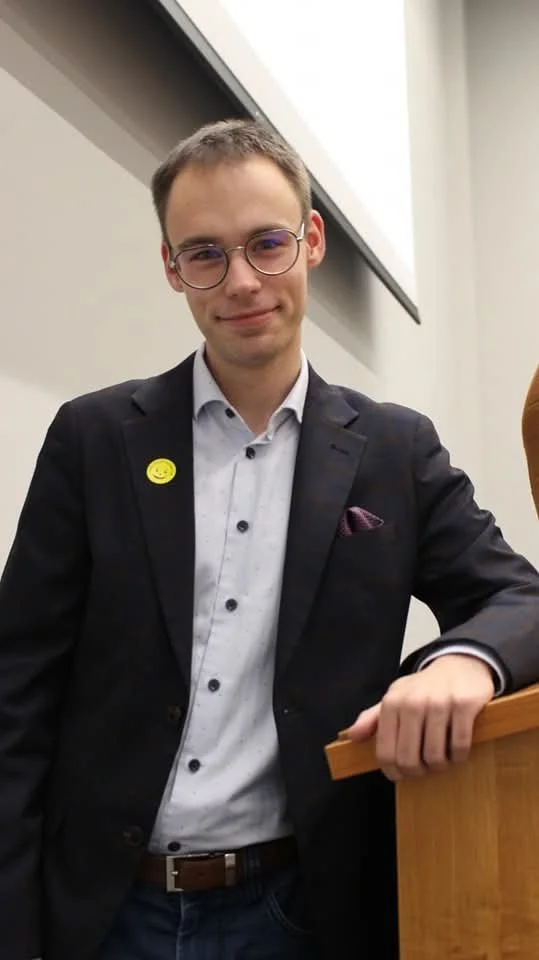 Young man with glasses, wearing a suit and white shirt, standing in front of a classroom or lecture hall, leaning on a wooden surface.