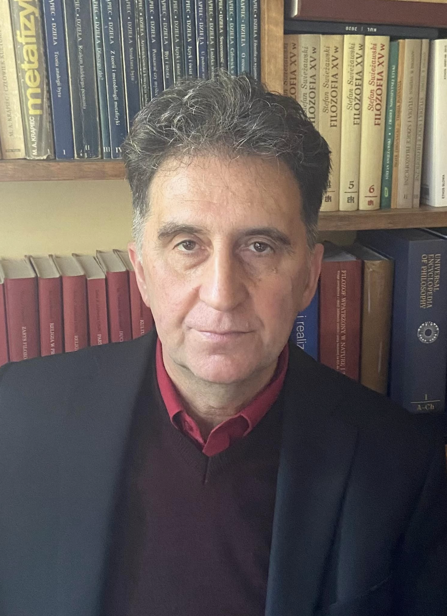 A middle-aged man with dark, graying hair, wearing a black blazer and red shirt, standing in front of a bookshelf filled with books on medicine, including titles about the human brain and medical dictionaries.