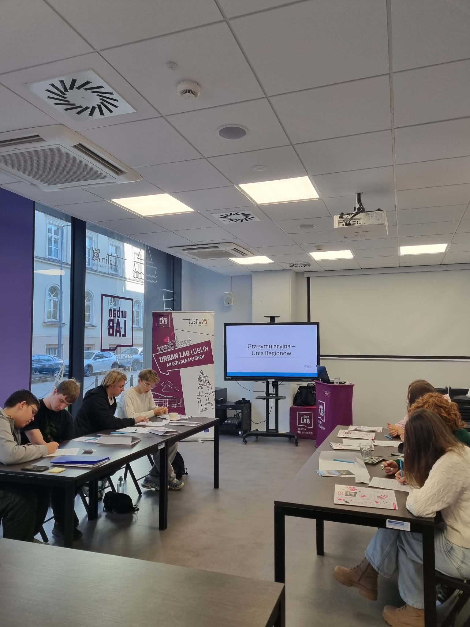 A group of people sitting at a long table in a conference room, engaging in a meeting or workshop. There is a large screen displaying text in Polish and various posters and materials on the table and walls, with windows showing buildings outside.